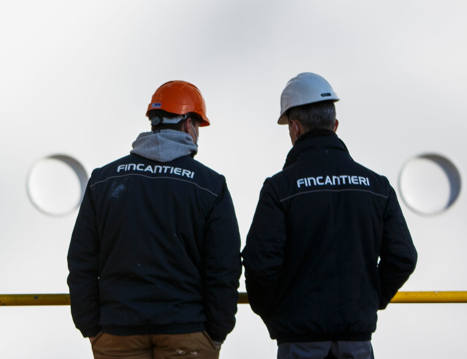 Fincantieri | Persone - Diversità, Equità e Inclusione: Due uomini con caschi e giacche Fincantieri vicino alla fiancata di una nave, grandi finestre rettangolari e superficie bianca, scena di osservazione o confronto in contesto industriale marittimo.