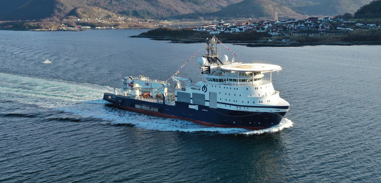 Prodotto - Offshore Tug/Supply Island Victory: Vista laterale della nave operativa in navigazione vicino a costa montuosa innevata, design moderno, antenne e attrezzature visibili sul ponte, edifici sparsi lungo la riva.