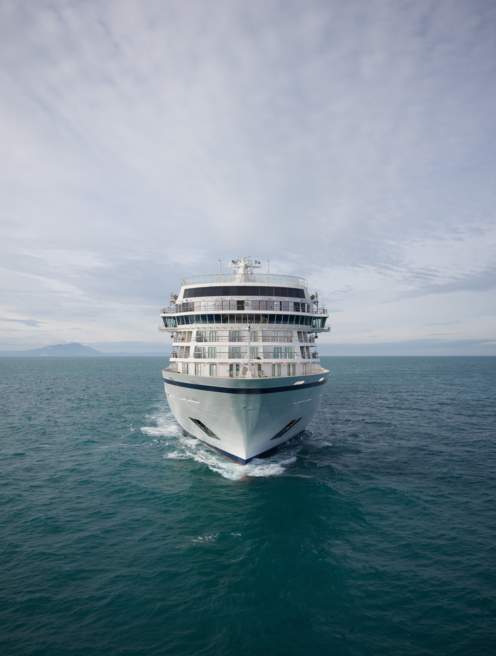 Fincantieri | Group - Business - Products - Cruise Ships - Viking Neptune: White cruise ship in operation, distant front view advancing in open sea, thin wake on water, wide cloudy sky above horizon.