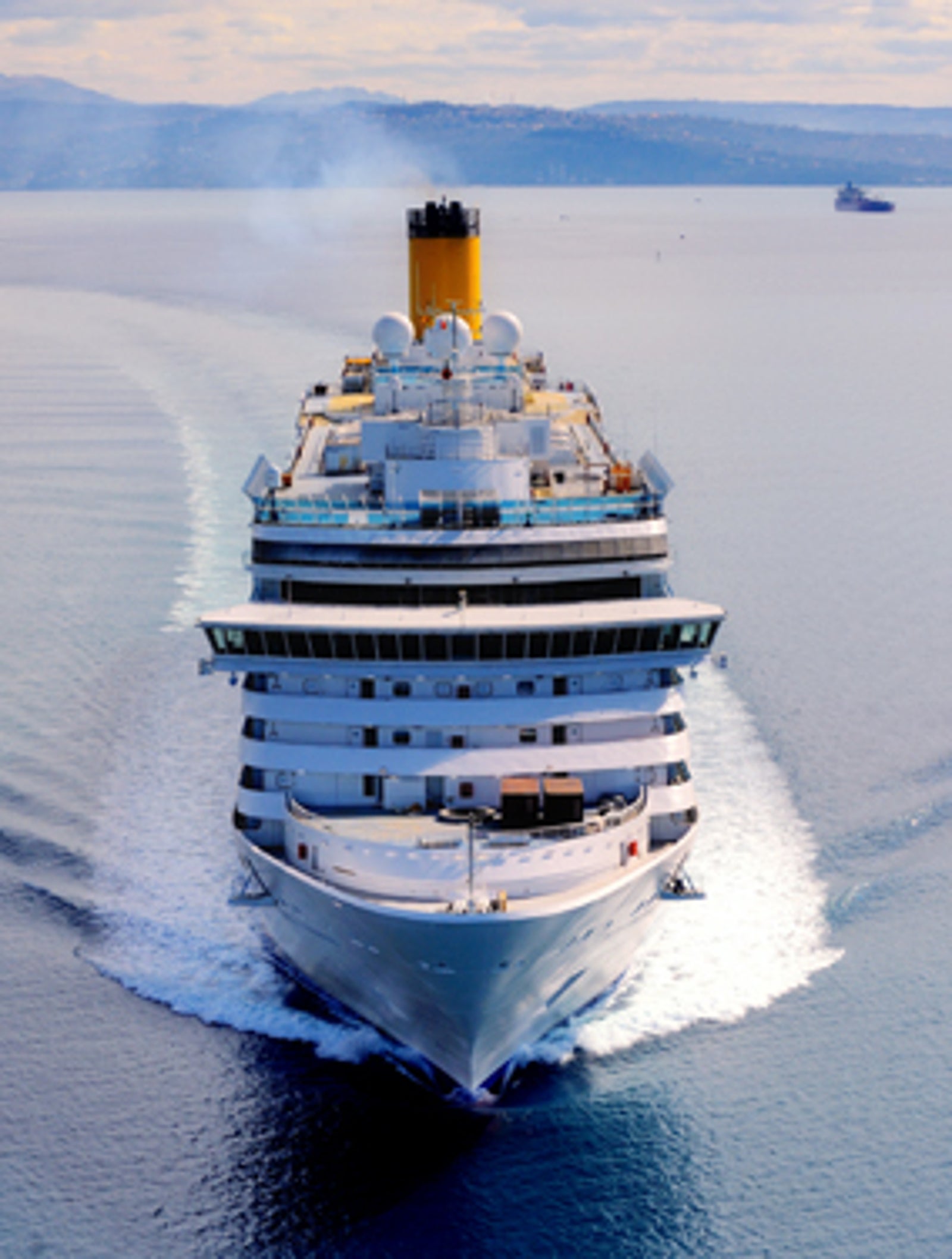Fincantieri | Gruppo - Business - Prodotti - Navi da Crociera - Costa Luminosa: Grande nave da crociera bianca operativa che naviga su acque calme e aperte sotto cielo parzialmente nuvoloso, scia visibile, colline distanti sull’orizzonte, scena che comunica viaggio e tranquillità.