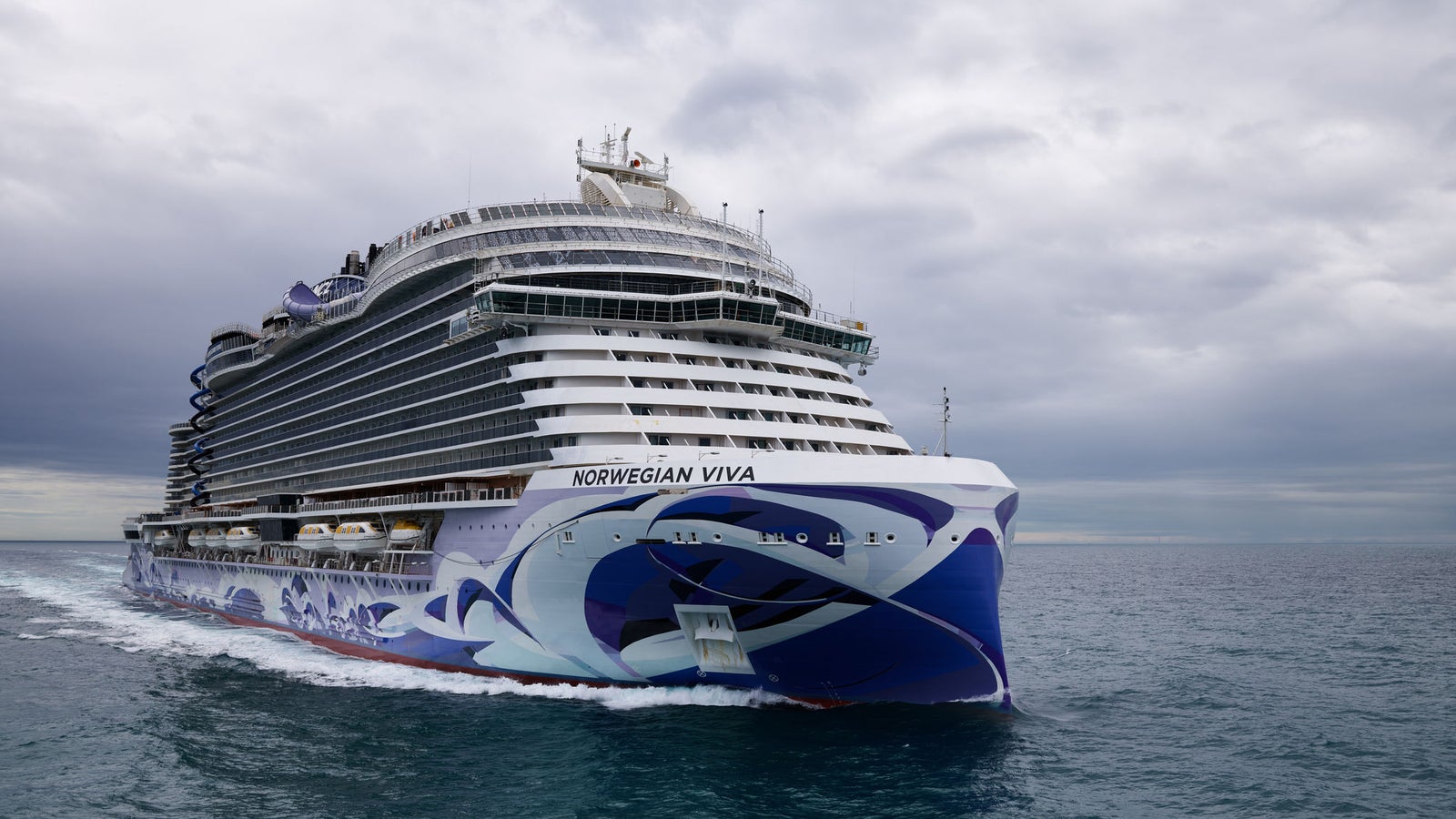  Fincantieri | Gruppo - Business - Prodotti - Navi da Crociera - Norwegian Viva: Nave da crociera Norwegian Viva operativa in navigazione su acque aperte calme sotto cielo nuvoloso, scafo con audace design a onde blu e bianco, nome 'Norwegian Viva' visibile a prua, più ponti e architettura moderna.