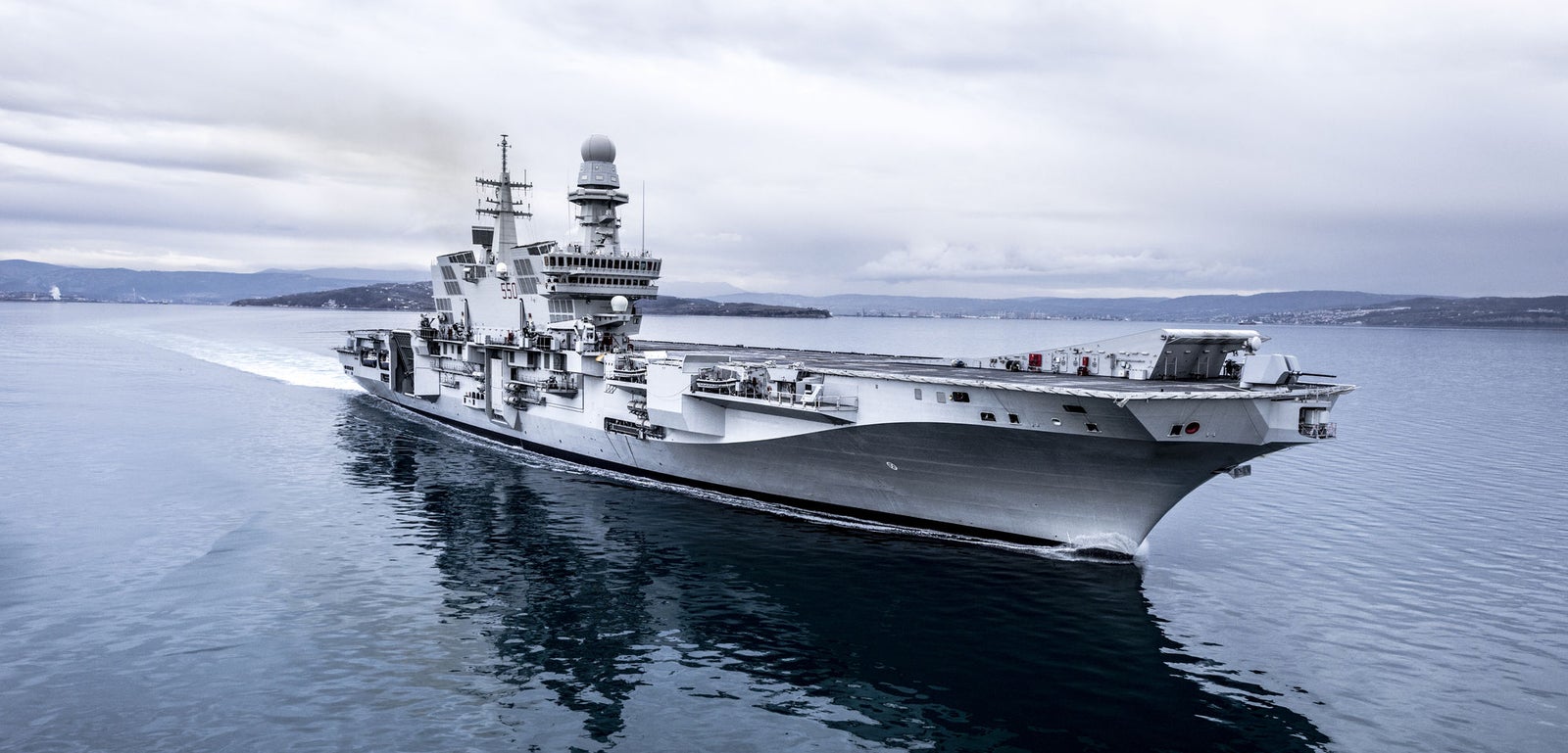 Fincantieri | Group - Business - Products - Naval Vessels - Aircraft Carriers and Amphibious Ships - Cavour: Large aircraft carrier "Cavour" in operation, starboard and front view, visible wake, sailing on calm dark water, overcast sky.