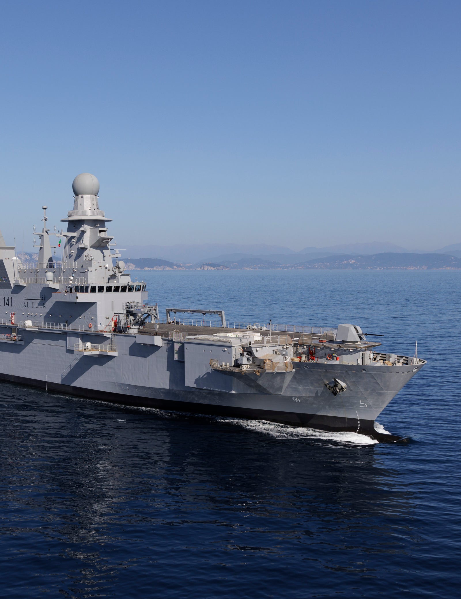  Fincantieri | Gruppo - Business - Prodotti - Navi Militari - Portaerei e Navi Anfibi - AL-FULK: Grande nave militare moderna in operazione, dotata di radar avanzati e design elegante, in navigazione su acque calme e aperte, cielo blu limpido, terra lontana sull’orizzonte.