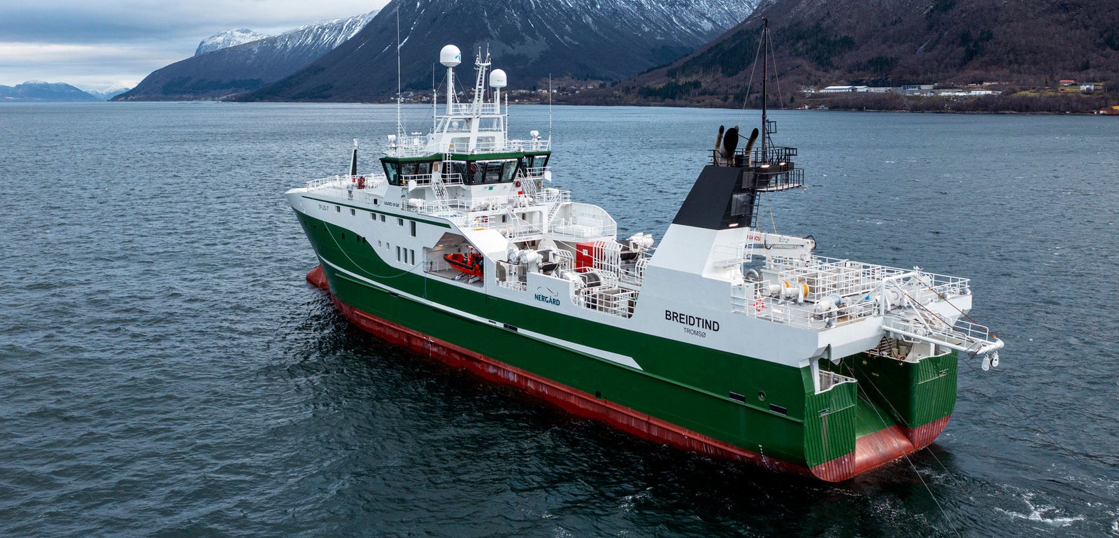 Prodotto - Peschereccio Bresting: Vista laterale della nave operativa su acque calme, scafo verde e bianco con nome Bresting visibile, dettagli strutturali evidenti, sfondo con montagne innevate e cielo nuvoloso.