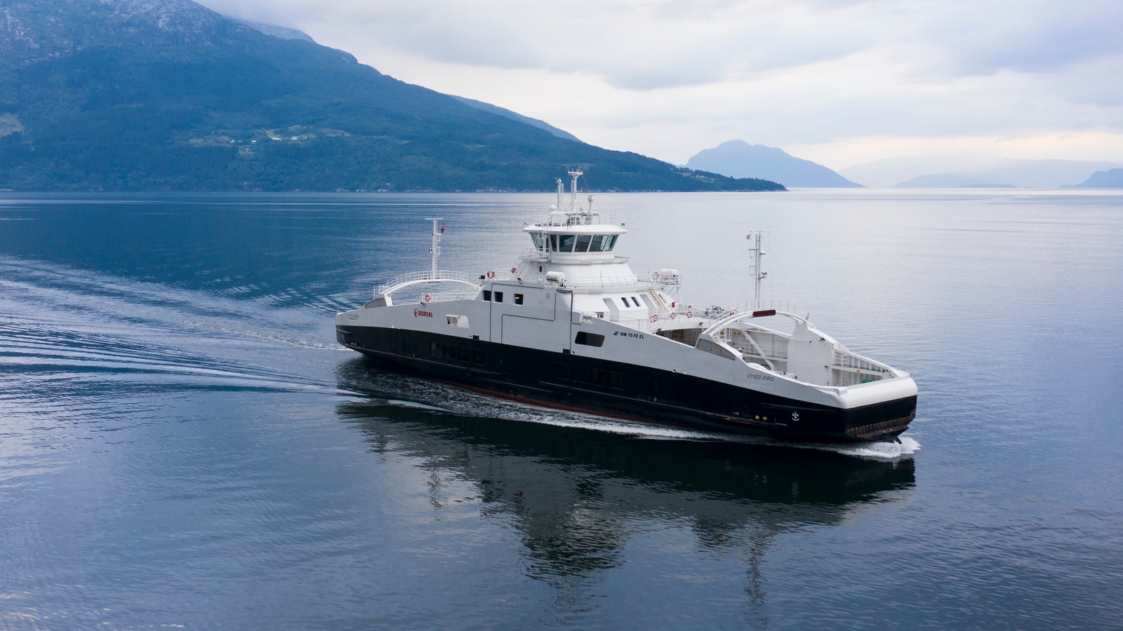 Fincantieri | Group - Business - Products - Ferries - Utnefjord: Modern Utnefjord ferry in operation, sleek design, navigating calm waters, surrounded by mountains, cloudy sky, ferry reflection visible on water.