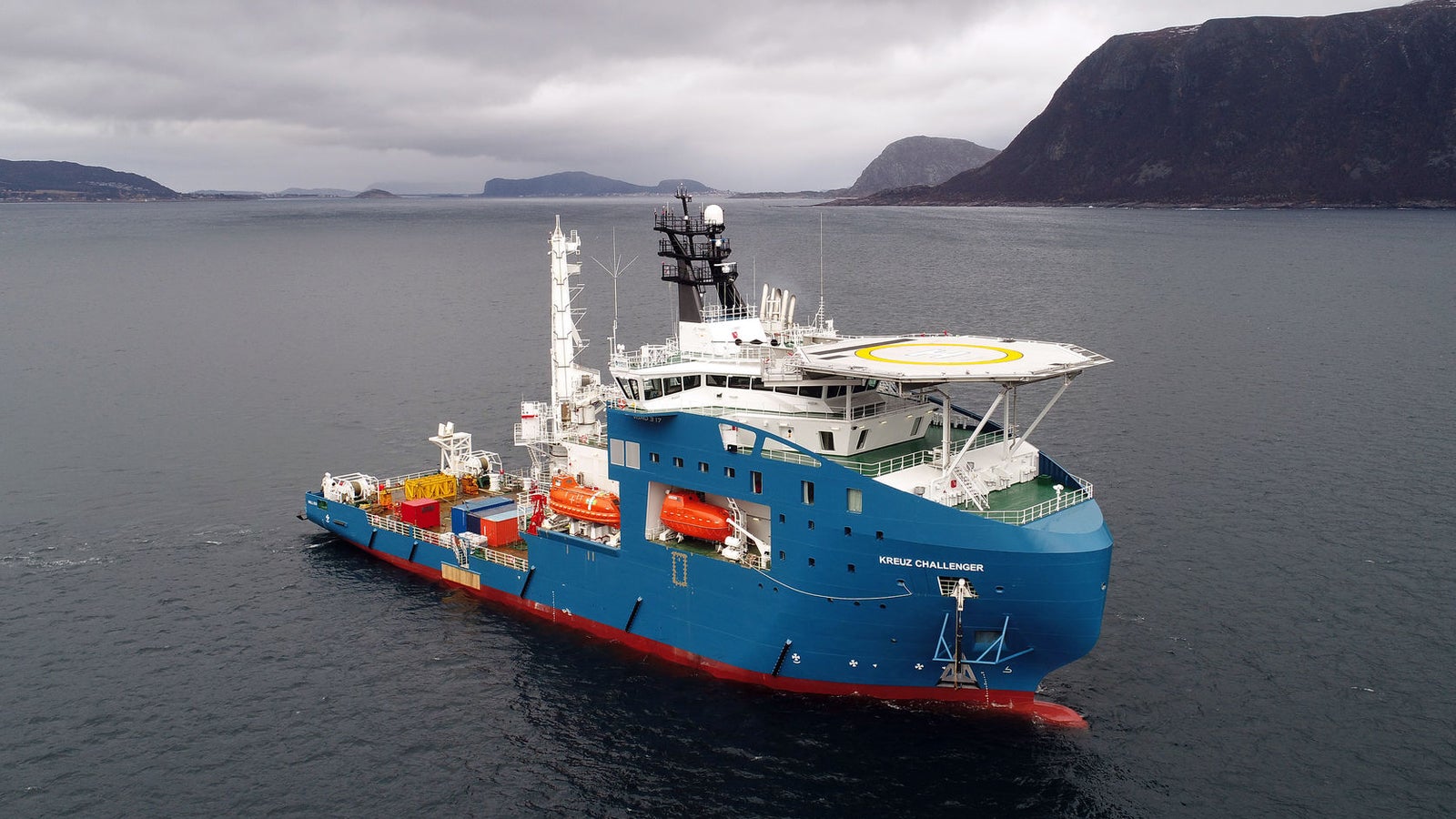 rodotto - Offshore Support Vessel Kreuz Challenger: Vista laterale della nave operativa in navigazione in acque calme tra fiordi, scafo blu, elisuperficie, gru e attrezzature arancioni sul ponte, nome Normand Installer chiaramente visibile.