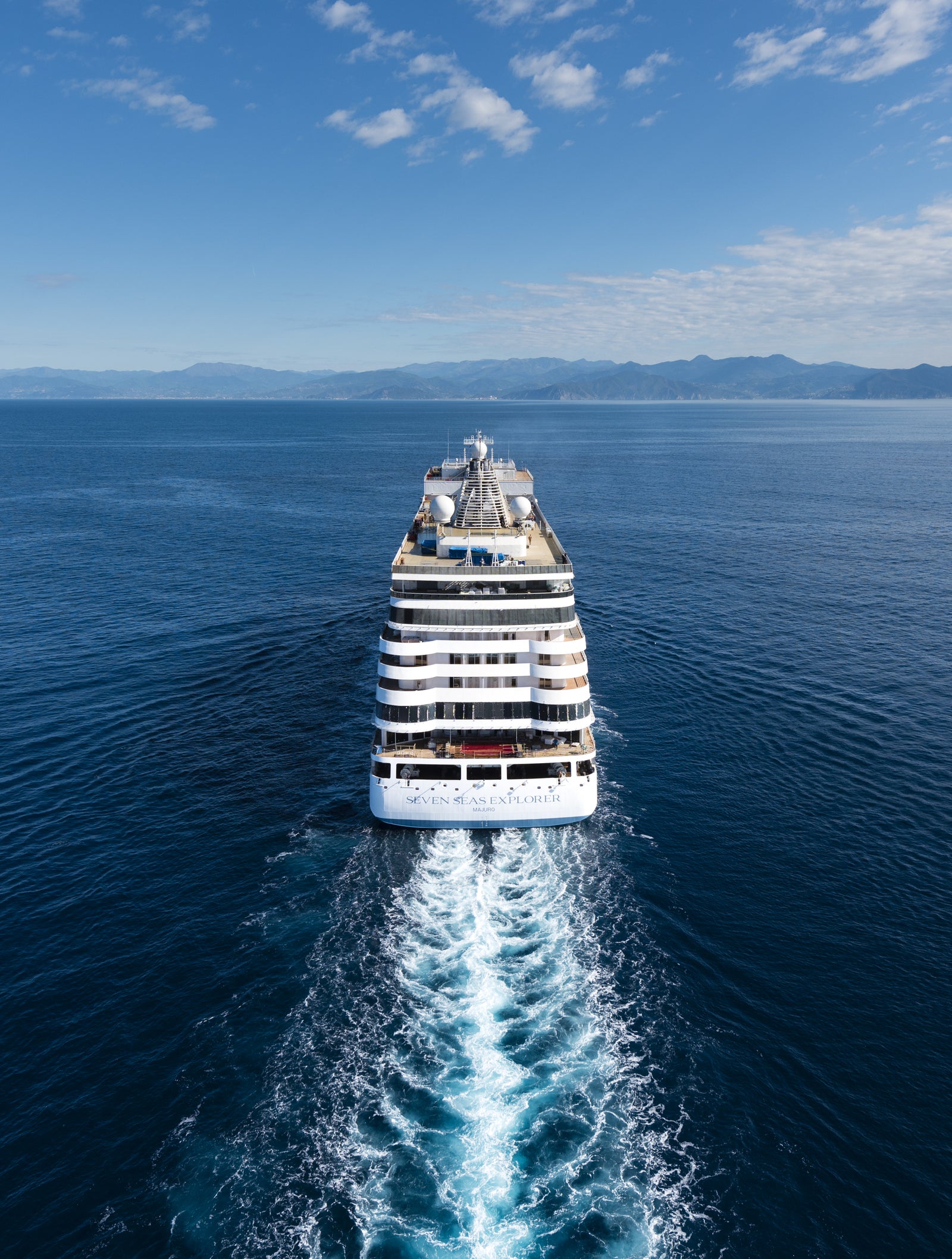  Fincantieri | Gruppo - Business - Prodotti - Navi da Crociera - Seven Seas Explorer: Grande nave bianca di lusso operativa con molteplici ponti e scialuppe di salvataggio visibili, naviga su acque calme sotto cielo sereno, scia dietro la nave e terre distanti all’orizzonte, ambiente marittimo tranquillo e suggestivo.