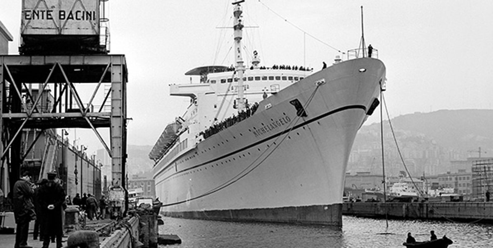 Fincantieri | About Us - Historic Ships: Large cruise ship docked in a crowded port, crowd visible on the deck, industrial structures and cranes with the writing 'ENTE BACINI', calm water, black and white photo highlighting the elegant design and vibrancy of the port.