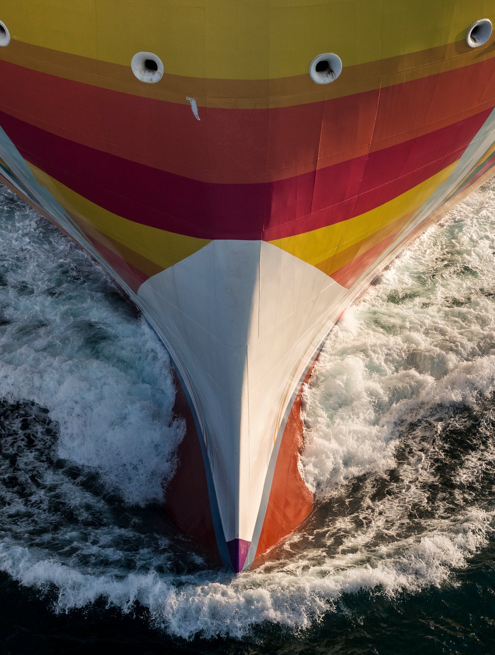 Fincantieri | Group - Business - Products - Cruise Ships - Norwegian Aqua: View of vibrant bow of large operational ship with yellow, red and orange striped hull, dynamic movement through waves in open sea.