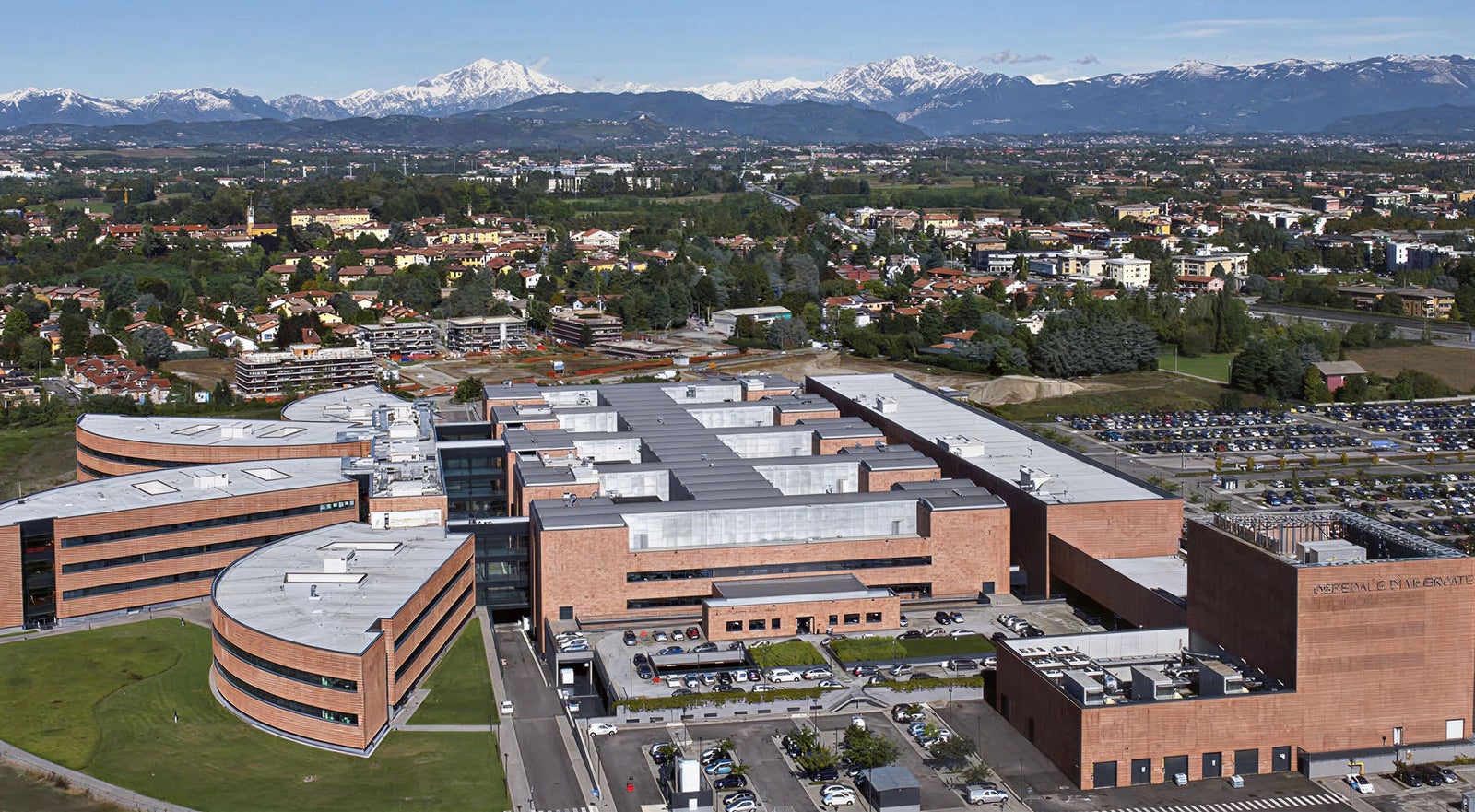 Fincantieri | Gruppo - Società - Controllate e collegate - Finso: Campus moderno esteso con edifici in mattoni e strutture architettoniche curve, circondato da aree verdi e parcheggi; contesto suburbano con montagne innevate in lontananza sotto cielo blu, enfasi su scala e disposizione del complesso in un ambiente suggestivo.