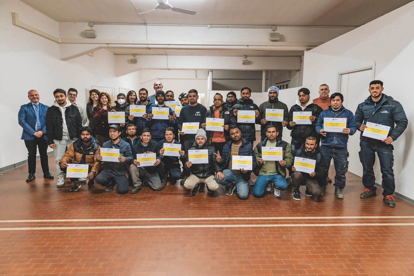 Newsroom - Comunicati stampa - 2025 - Gruppo Fincantieri: Fondazione Fincantieri e Società Dante Alighieri: ad Ancona la consegna degli attestati dei corsi di lingua italiana per lavoratori stranieri: Foto di gruppo con corsisti e lavoratori stranieri che mostrano i certificati di lingua italiana ricevuti, insieme ai rappresentanti di Fondazione Fincantieri e Società Dante Alighieri in un ambiente interno luminoso.