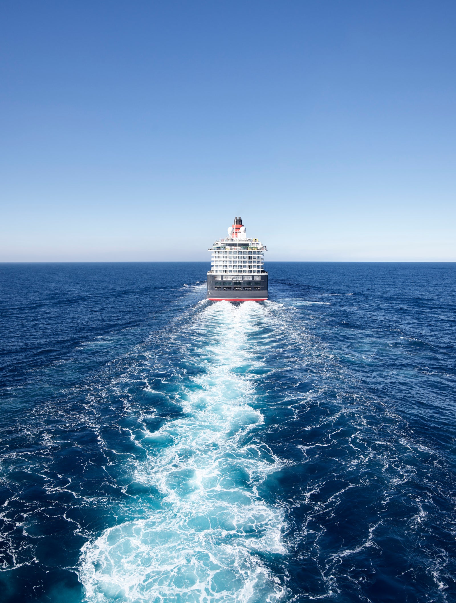Fincantieri | Group - Business - Products - Cruise Ships - Queen Anne: Cruise ship stern rear view in operation, sailing away on deep blue ocean, white foamy wake spreading symmetrically, clear sky.