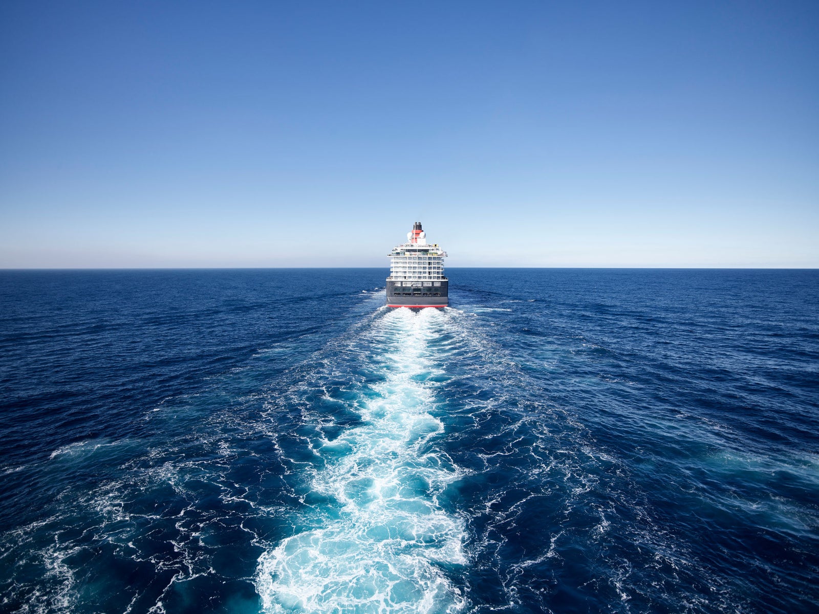 Fincantieri | Group - Business - Products - Cruise Ships - Queen Anne: Cruise ship stern rear view in operation, sailing away on deep blue ocean, white foamy wake spreading symmetrically, clear sky.
