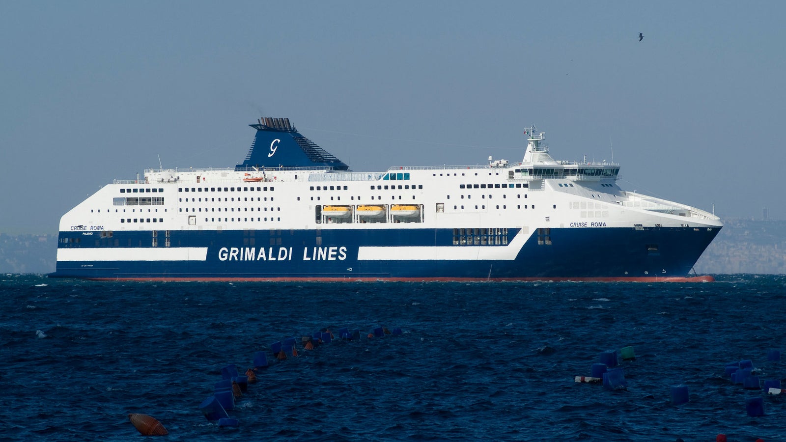 Fincantieri | Group - Business - Products - Ferries - Cruise Barcellona: Large Grimaldi Lines ferry cruising through open waters on a clear day; white and blue color scheme with 'Grimaldi Lines' text visible on side; calm seas and distant land in background; ferry design indicates passenger and cargo transport.