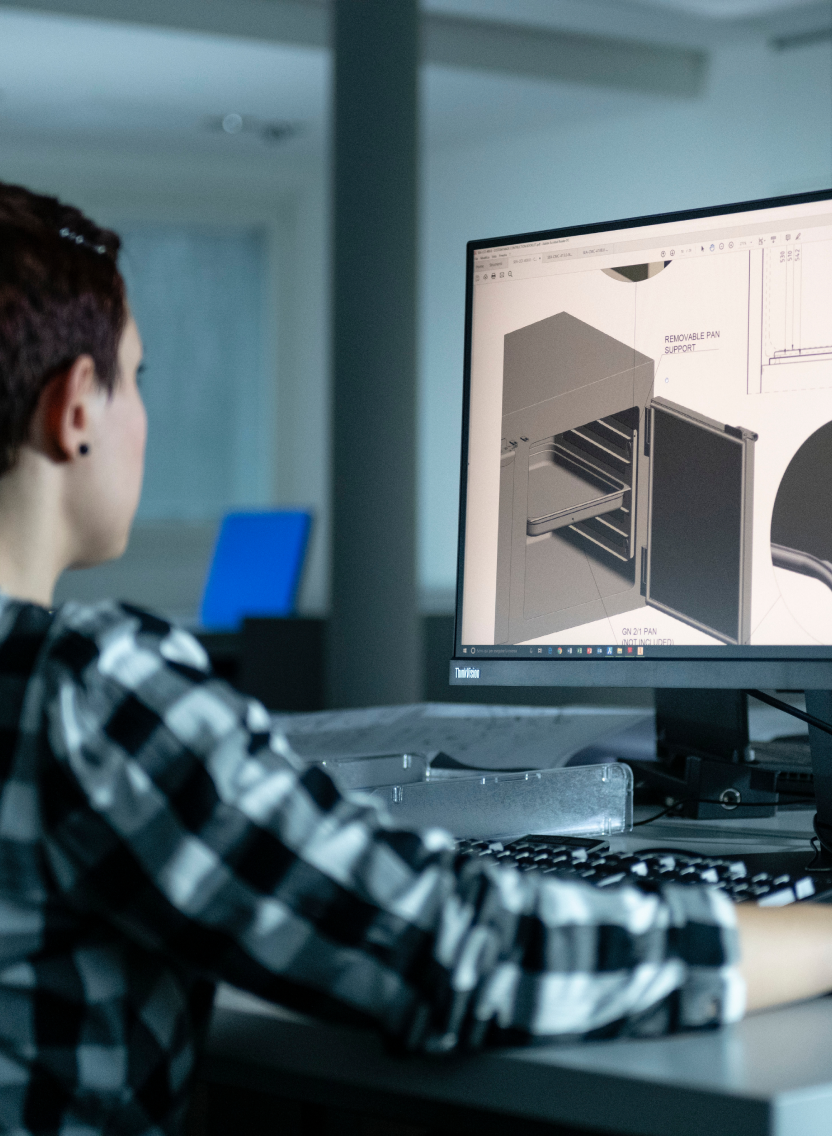 Fincantieri | People - Living Fincantieri - Diversity, Equity and Inclusion - Stories and Testimonials: A person is seated at a desk, working on a computer displaying a 3D model design. The setting appears to be a modern office environment with minimalistic decor. The individual is wearing a checkered shirt, and the screen shows detailed technical drawings and measurements