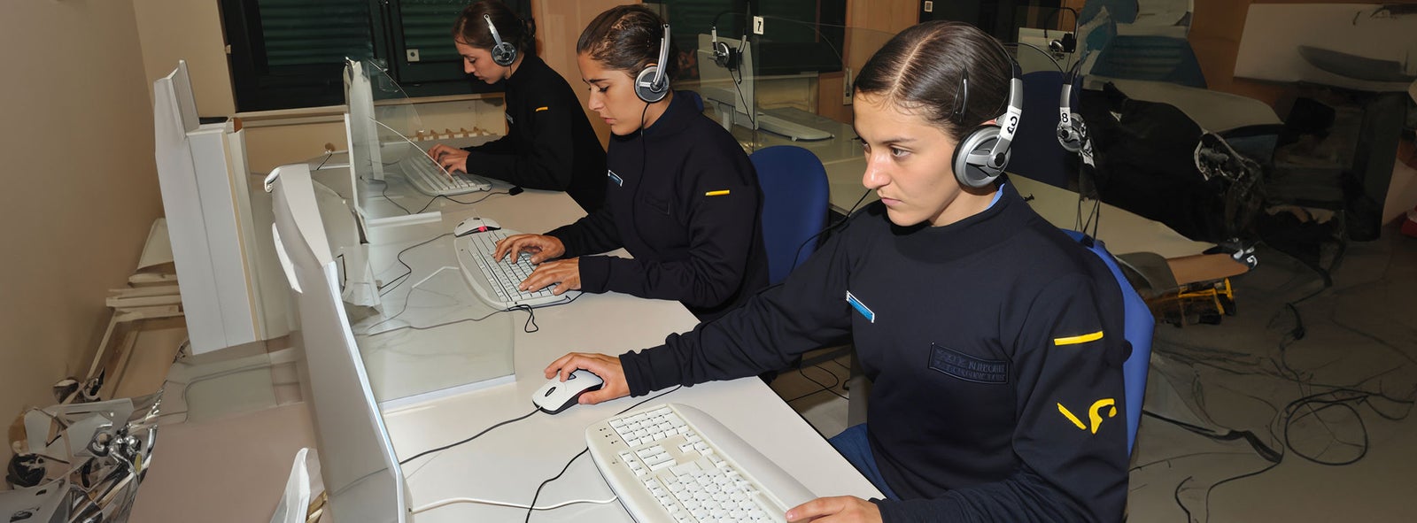 Fincantieri | Business Services - After-Sales Services - Training & Support: Three individuals are seated at computer workstations in a professional setting, wearing matching dark uniforms and headsets. The environment appears to be an office or training facility with glass partitions and organized desks. The uniforms feature visible yellow and blue accents, suggesting a coordinated team or organization.