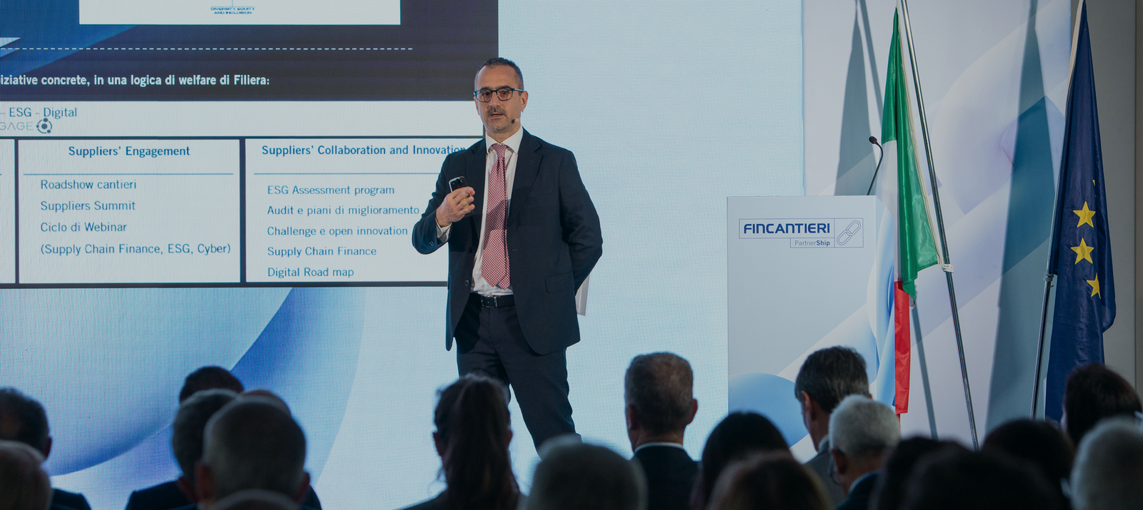 Fornitori e Partner – Fincantieri | Partnership: Relatore professionale presenta a un evento aziendale, grande schermo con slide contenente termini come “ESG Digital” e “Supply Chain Finance, ESG, Open”, pubblico in abiti formali, bandiere italiana e UE visibili, focus su collaborazione e innovazione business.