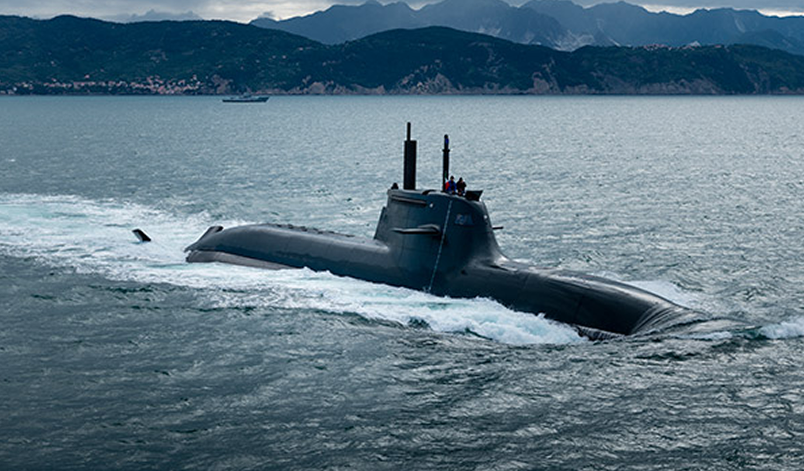 Fincantieri | About Us - Heritage and Future: Sleek, functional military submarine with visible antennas and periscopes, sailing partially submerged on calm waters, with a mountainous coastal background and cloudy sky.