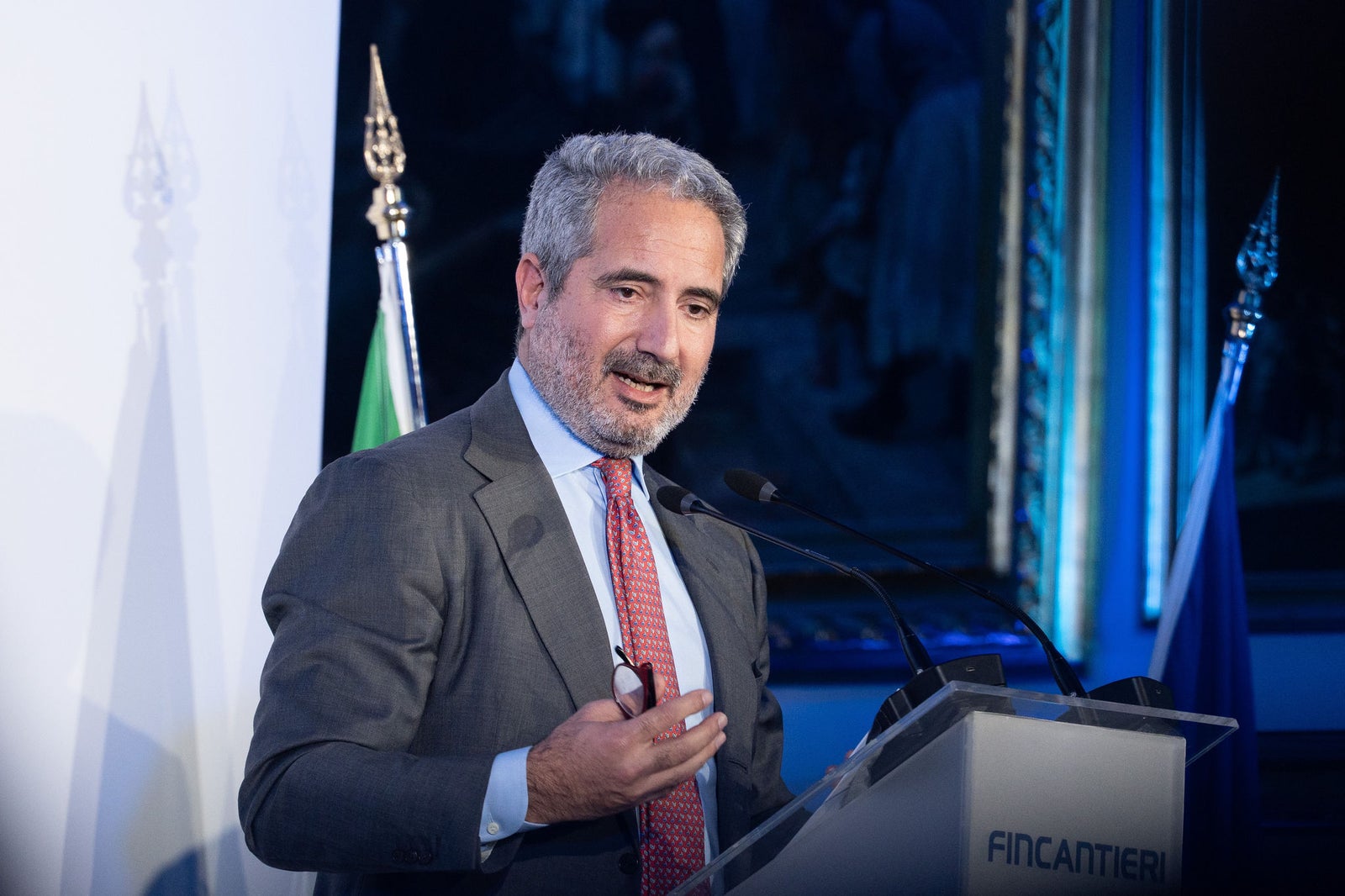 Newsroom - Comunicati stampa - 2025 - Gruppo Fincantieri: Un anno di Fondazione Fincantieri: storia, innovazione e impatto sociale: Primo piano dello speaker istituzionale durante l’evento annuale della Fondazione Fincantieri, a un podio con logo, bandiere italiana ed europea sullo sfondo, in sala conferenze illuminata di blu.