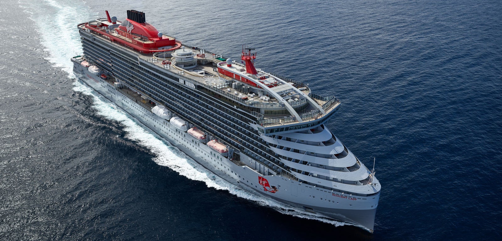Fincantieri | Group - Business - Products - Cruise Ships - Resilient Lady: Large Virgin Voyages operational cruise ship with modern design and red details, multiple decks, name 'Resilient Lady' visible on hull, distinctive red funnel, sailing in calm waters under partly cloudy sky, atmosphere of luxury and maritime adventure.