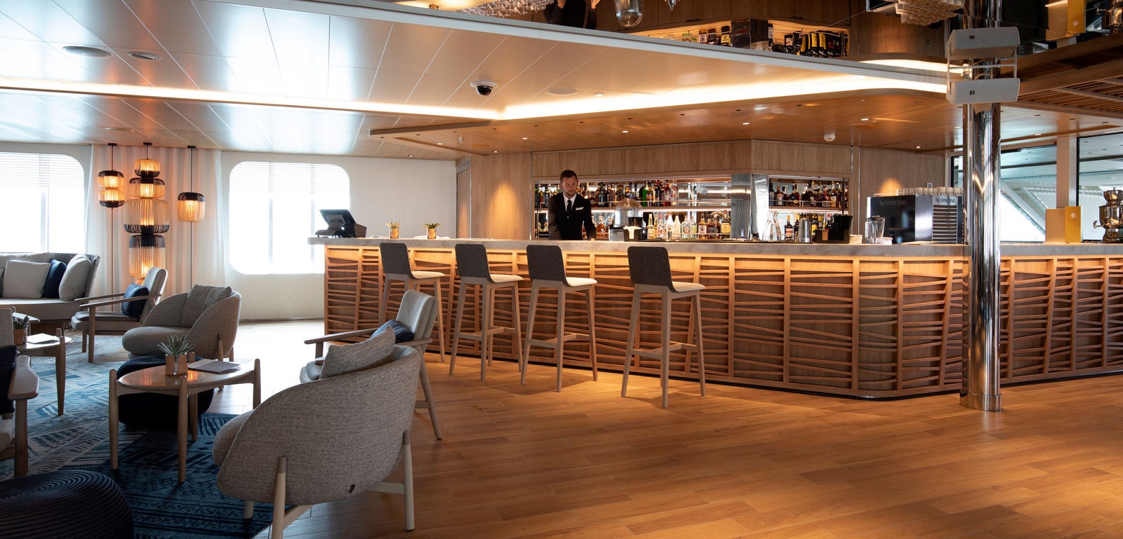 Fincantieri | Gruppo - Business - Prodotti - Navi da Crociera - Le Lapérouse: Lounge bar elegante e contemporaneo con accenti in legno, arredi minimalisti, bartender dietro bancone assortito, disposizioni di seduta confortevoli, illuminazione calda e superfici riflettenti, atmosfera raffinata.