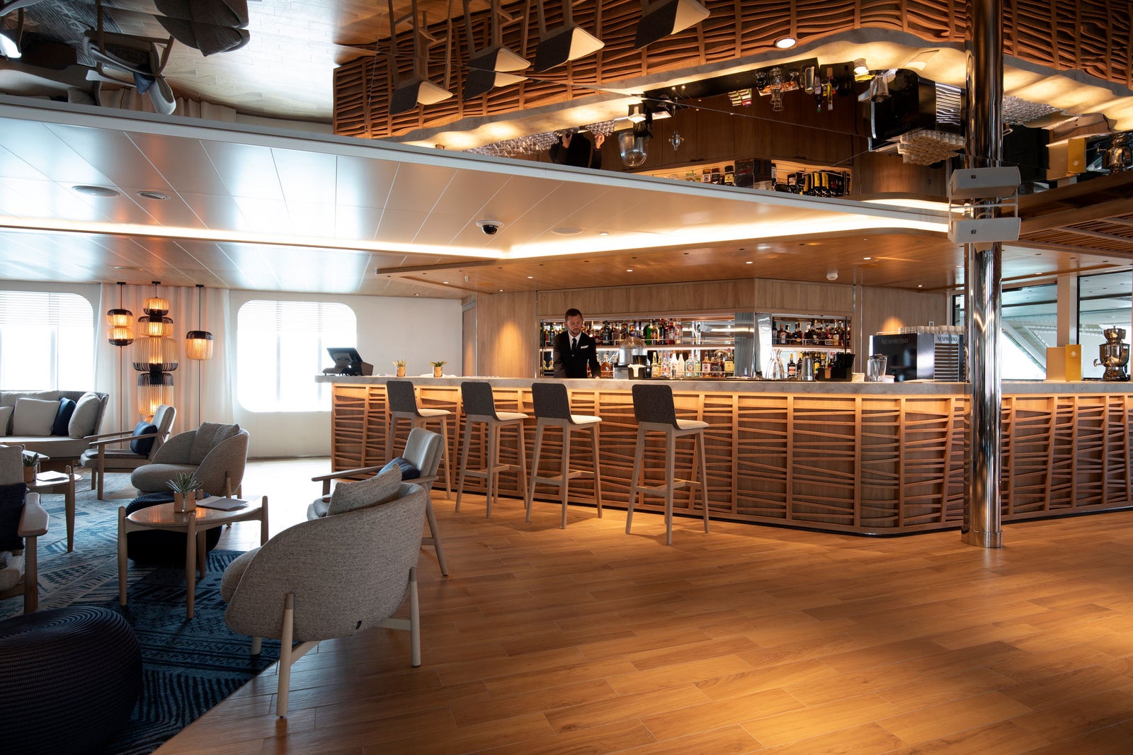 Fincantieri | Gruppo - Business - Prodotti - Navi da Crociera - Le Lapérouse: Lounge bar elegante e contemporaneo con accenti in legno, arredi minimalisti, bartender dietro bancone assortito, disposizioni di seduta confortevoli, illuminazione calda e superfici riflettenti, atmosfera raffinata.