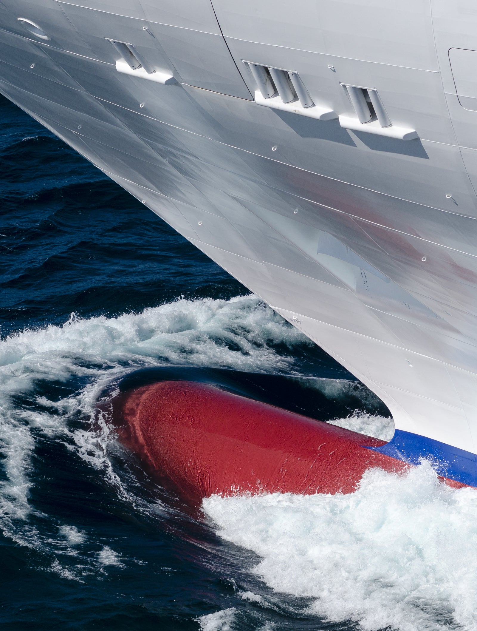 Fincantieri | Group - Business - Products - Cruise Ships - Costa Fascinosa: Detailed view of ship’s bow in operation slicing through ocean waves, red bulbous bow visible beneath waterline, white exterior contrasts with deep blue sea, sleek design, image captures dynamic movement and maritime engineering.
