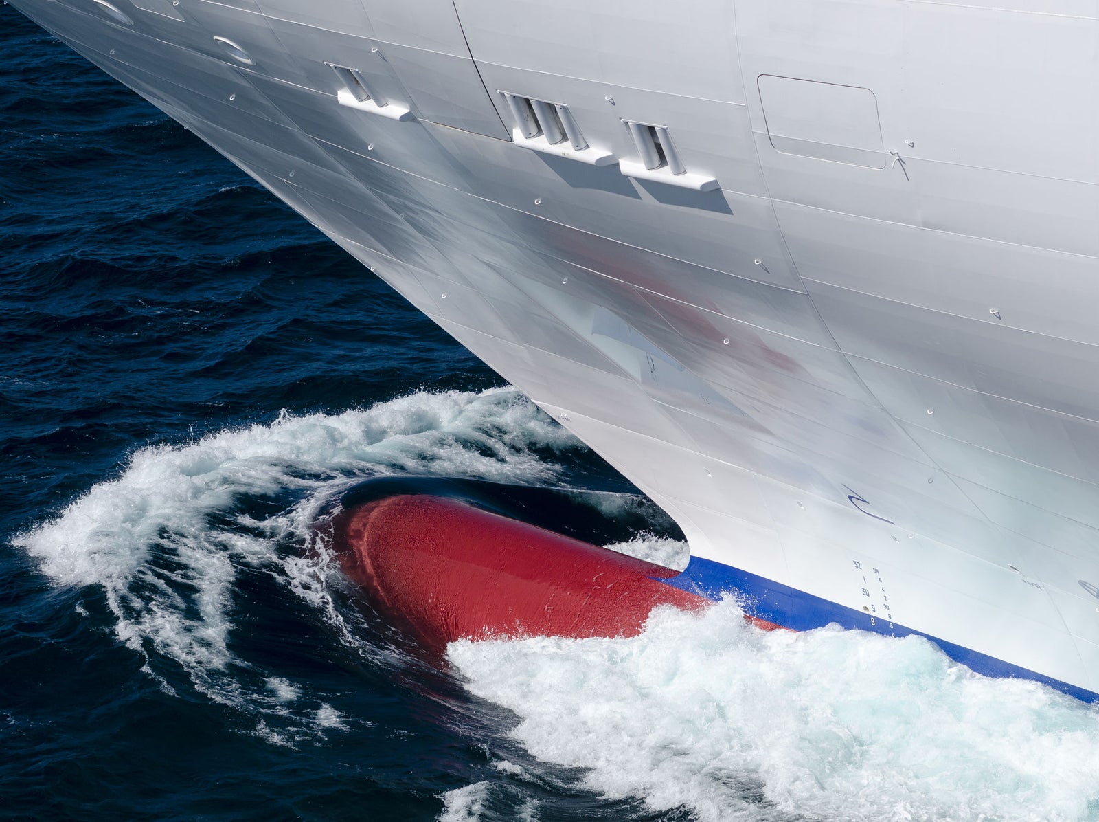  Fincantieri | Group - Business - Products - Cruise Ships - Costa Fascinosa: Detailed view of ship’s bow in operation slicing through ocean waves, red bulbous bow visible beneath waterline, white exterior contrasts with deep blue sea, sleek design, image captures dynamic movement and maritime engineering.