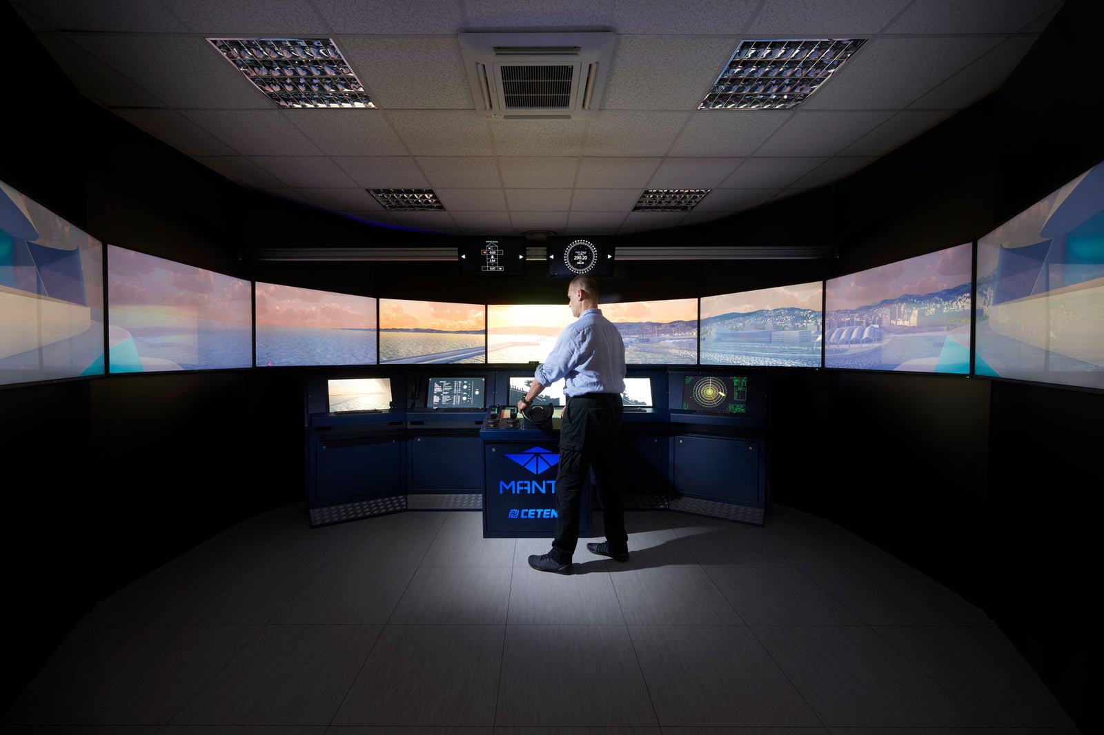 Fincantieri | Group - Subsidiaries and Affiliates - Fincantieri Cetena: Man stands in high-tech maritime simulation room, surrounded by multiple large screens displaying oceanic visuals; professional training or control setting; central console with 'MANTIS' and 'CETENA' text clearly visible; well-lit, modern, professional ambience.