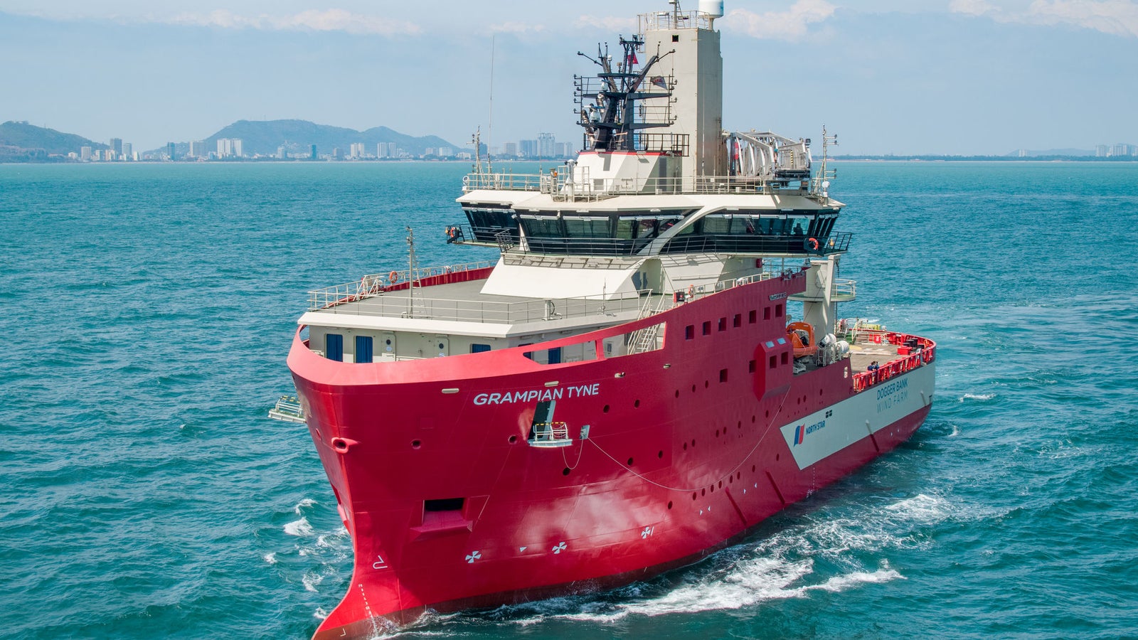 Prodotto - Offshore Support Vessel Grampian Tyne: Vista laterale della nave operativa in navigazione, scafo rosso, equipaggiamenti e strutture moderne sul ponte, nome Grampian Tyne visibile, sfondo urbano costiero e colline distanti.