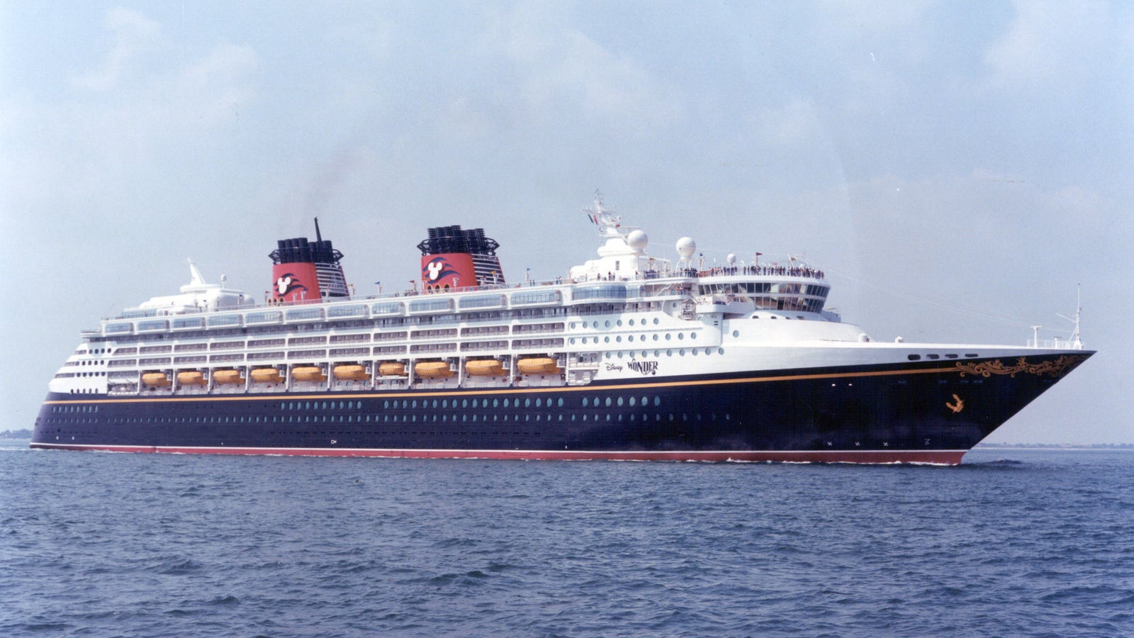 Fincantieri | Group - Business - Products - Cruise Ships - Disney Wonder: Side view of Disney Wonder cruise ship in operation navigating, sleek design, dual red and black funnels, multiple decks, visible lifeboats, navy blue hull with white details.