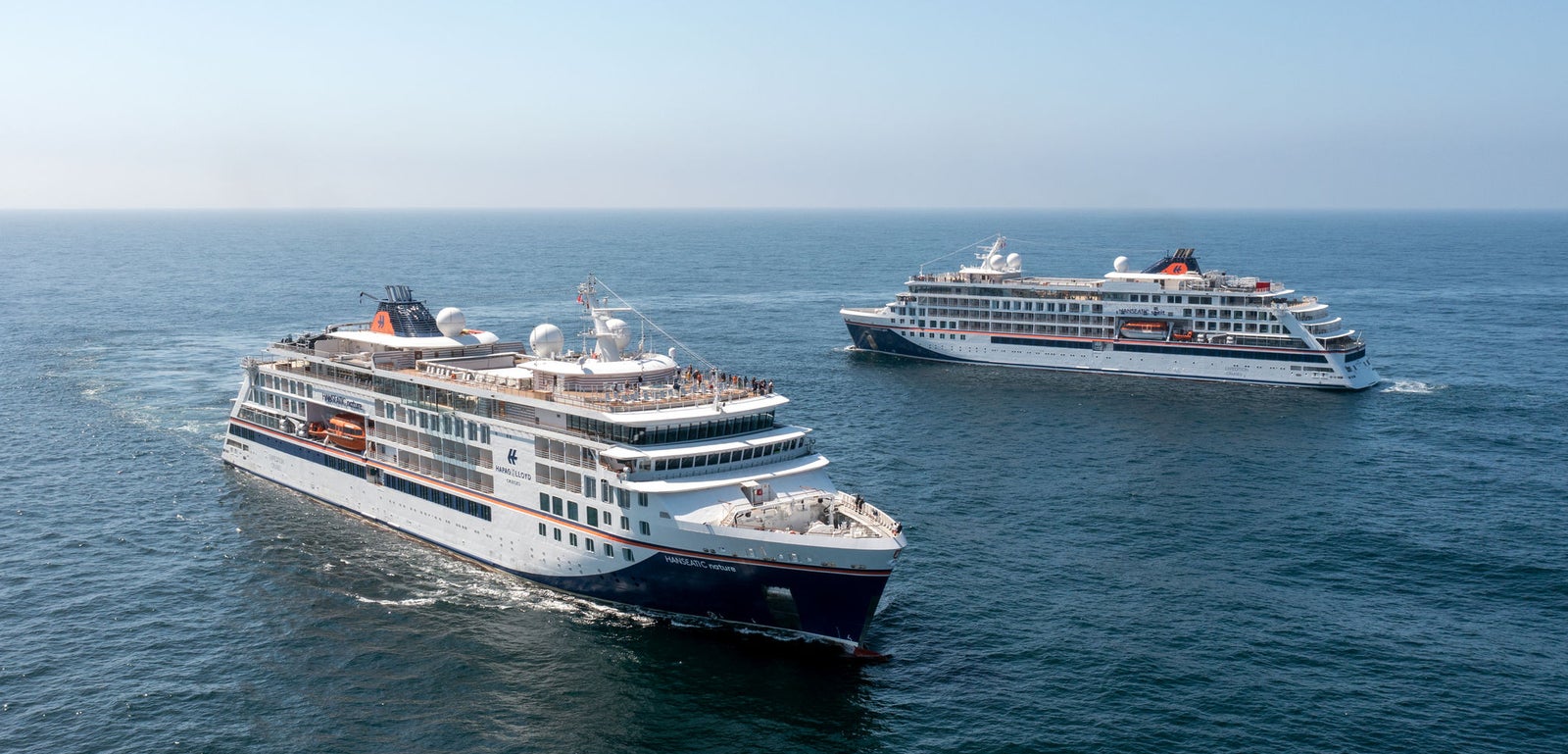 Fincantieri | Group - Business - Products - Cruise Ships - HANSEATIC spirit: Two modern operational cruise ships sailing parallel on calm open waters under clear blue sky, multiple decks with outdoor seating and observation areas, scene evokes serene maritime travel for leisure and exploration.