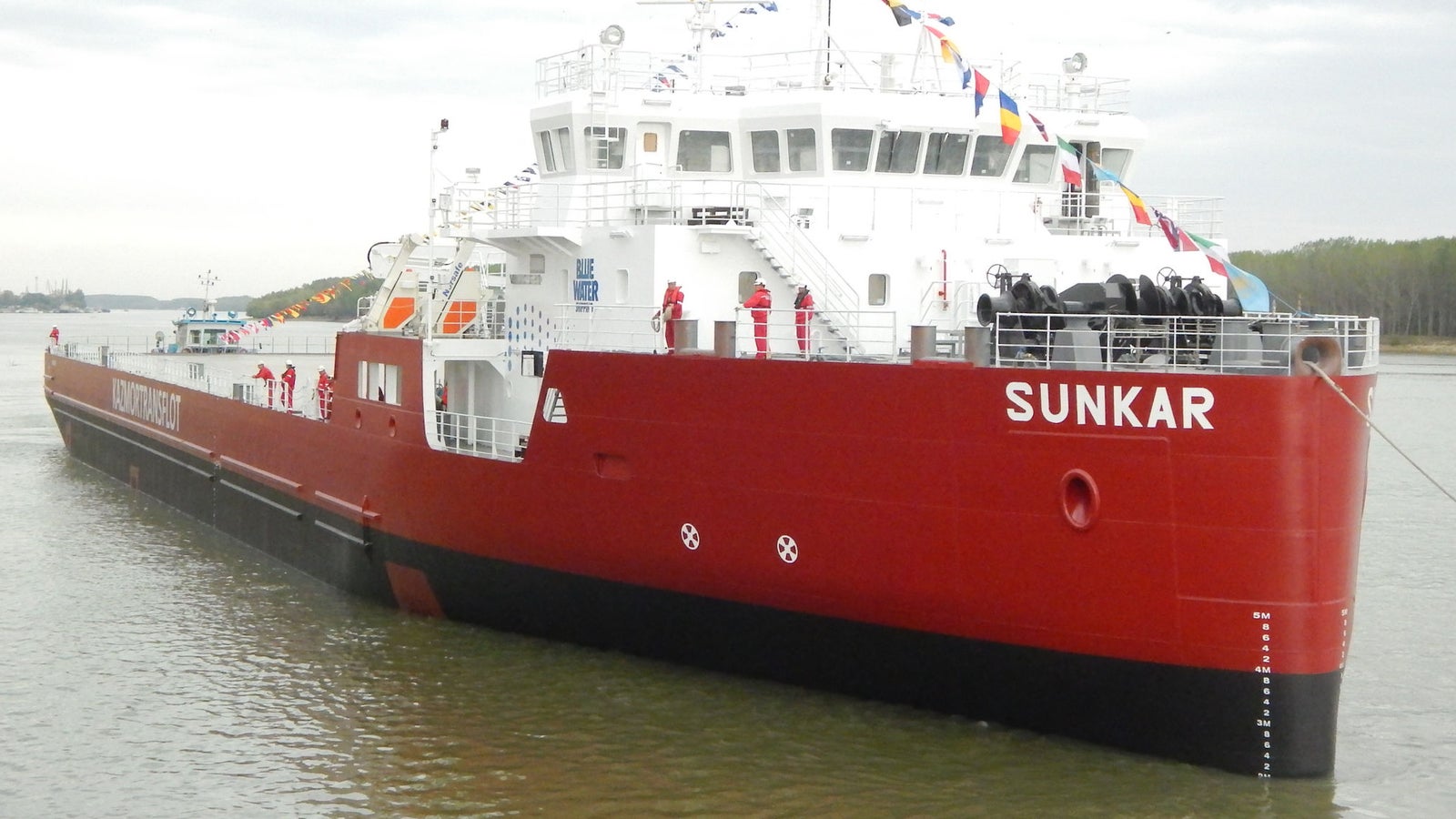 Prodotto - Nave Cargo Sunkar: Vista laterale della nave ormeggiata o in navigazione, scafo rosso con nome Sunkar visibile, bandiere colorate, presenza dell’equipaggio in uniforme rossa sul ponte, cielo nuvoloso.