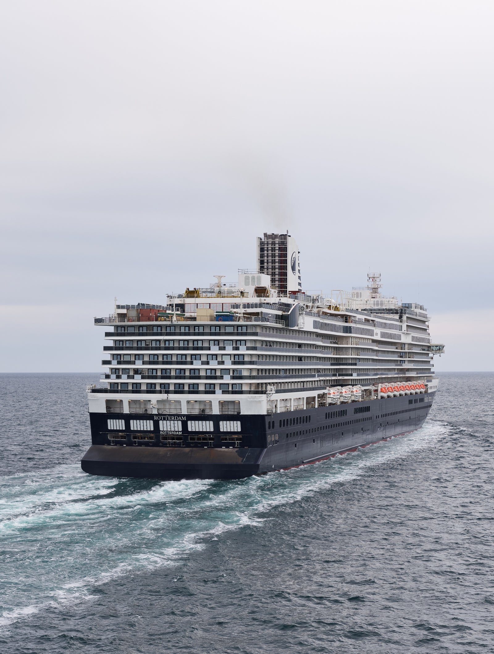 Fincantieri | Group - Business - Products - Cruise Ships - Rotterdam: Stern view of large operational cruise ship sailing on calm open waters under cloudy sky, multiple decks and visible funnels emitting light smoke.