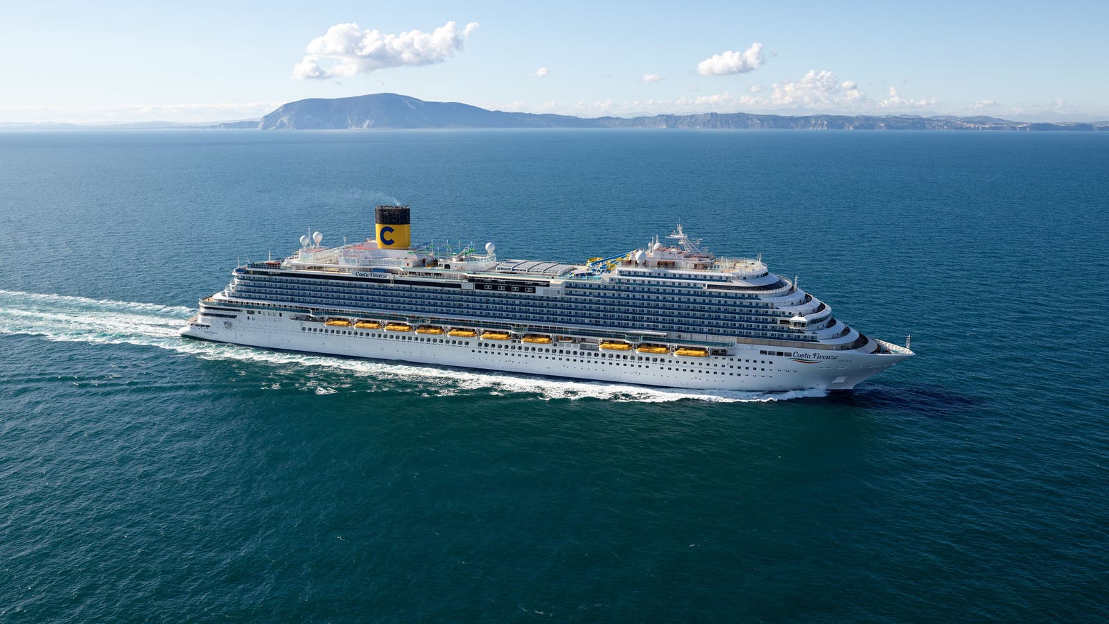 Fincantieri | Group - Business - Products - Cruise Ships - Costa Firenze: Large operational cruise ship side view, multiple decks and visible lifeboats, yellow funnel with 'C' logo, sailing on calm waters with mountainous coastline in background.