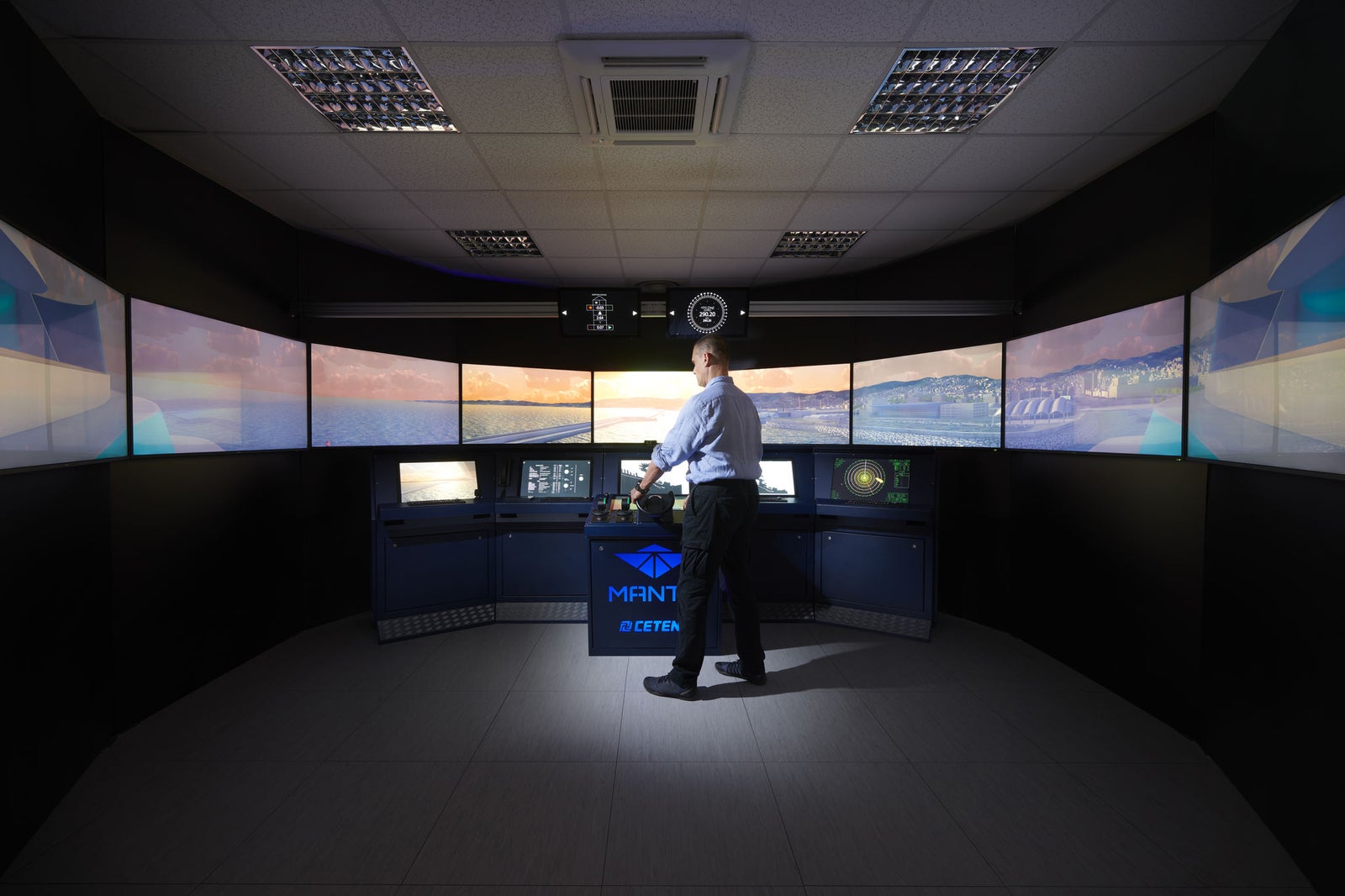 Fincantieri | Group - Business - Products - Cybersecurity - Simulation system (MANTIS): Man stands at center of high-tech maritime simulation system, surrounded by multiple large screens displaying nautical visuals; professional training or control room with dim lighting, 'MANTIS' and logo clearly visible on central console.