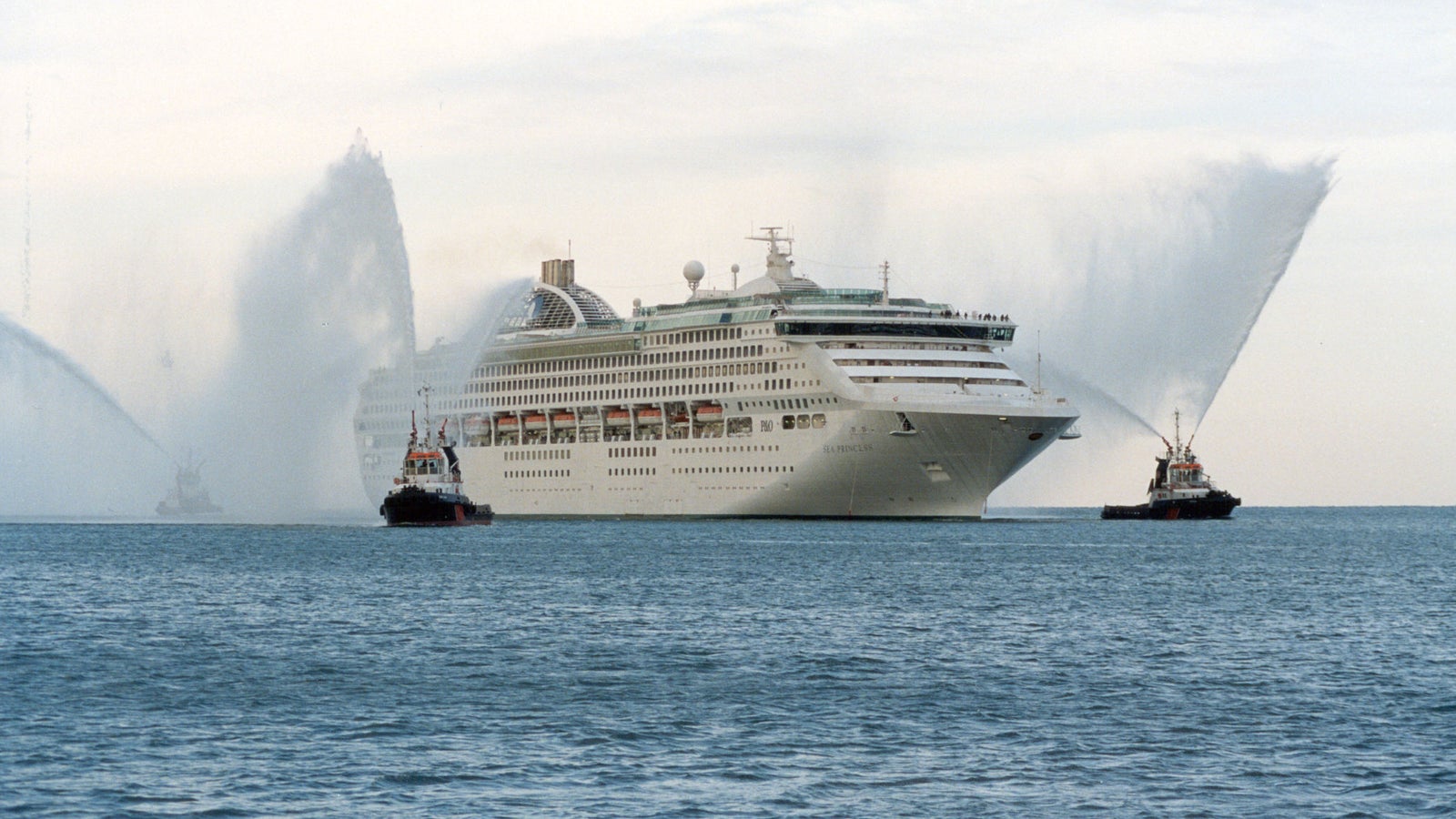 Fincantieri | Group - Business - Products - Cruise Ships - Sea Princess: Side view of Sea Princess cruise ship in operation navigating, multiple decks and large windows, accompanied by two tugboats with celebratory water jets.