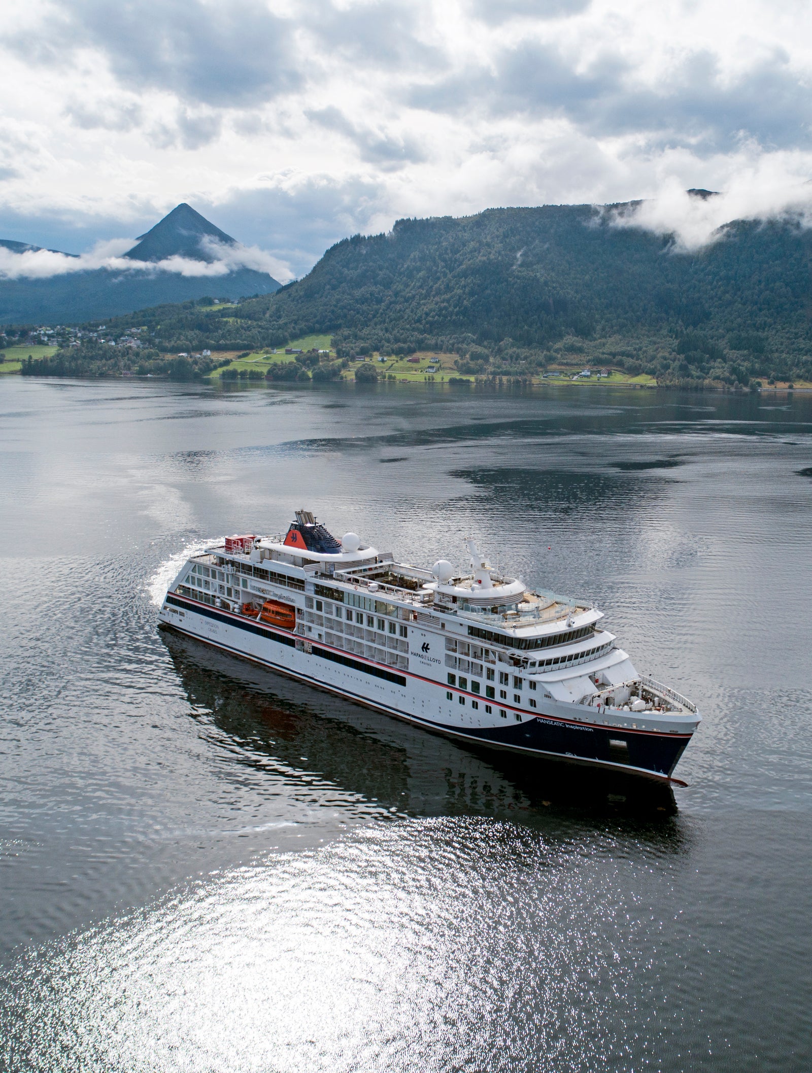 Prodotto - Nave da Crociera Hanseatic Inspiration: Grande nave da crociera naviga in acque calme tra spettacolari paesaggi di fiordo. Colline verdi lussureggianti, montagne nebbiose e acque riflettenti sotto cielo nuvoloso. Esterno bianco con scialuppe visibili ed elementi di design dettagliati. Nave operativa