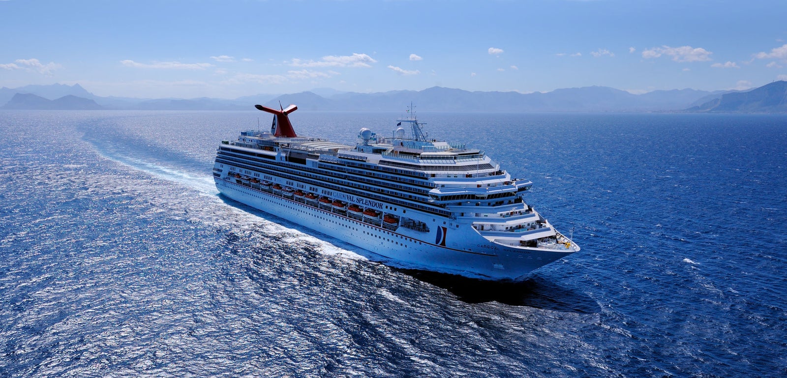 Fincantieri | Group - Business - Products - Cruise Ships - Carnival Splendor: Overhead and side view of large operational cruise ship gliding on vast sparkling blue ocean under clear sky, multiple decks, distinctive red funnel, distant landforms on horizon, serene setting evoking leisure and travel.