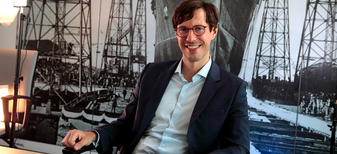 Fincantieri – People: Man in a navy blue suit and white shirt sitting at a desk in a professional office; in the background, a large black-and-white historical photograph of industrial structures and vessels, with modern lighting and a visible desk lamp.