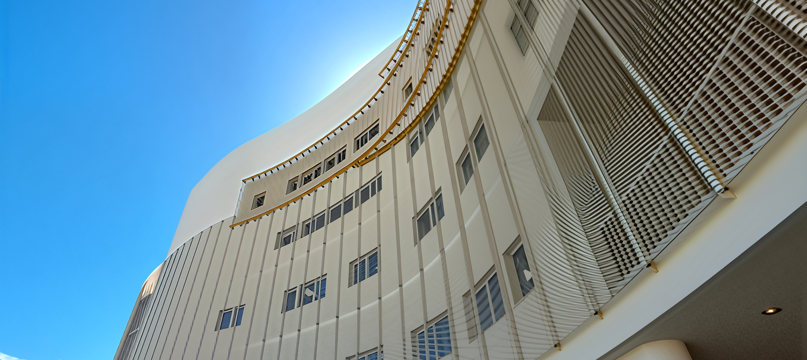 Fincantieri | Group - Business - Products - Maritime/Hydraulic Works - Contemporary building: Contemporary building with curved facade featuring vertical lines and multiple windows; set against bright blue sky for striking contrast, golden accents along edges add elegance to architectural design.