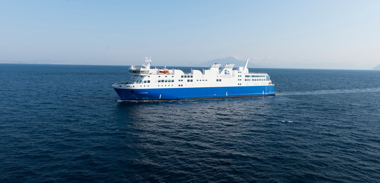 Fincantieri | Group - Business - Products - Ferries - Cruise FA Gauthier: White and blue "FA Gauthier" ferry in operation, multiple decks, visible lifeboats, cruising across calm open waters, clear blue sky.