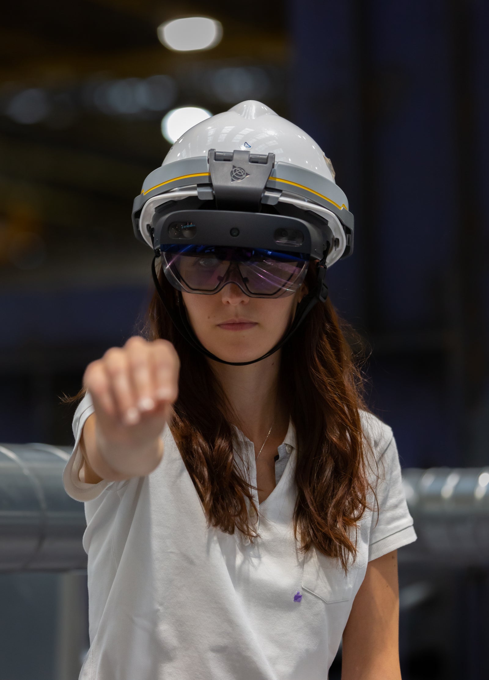 Fincantieri | About Us - Strategic Vision: Person wearing a white safety helmet and white polo shirt in an industrial environment with metal structures and soft lighting, making a forward gesture indicating action or communication.