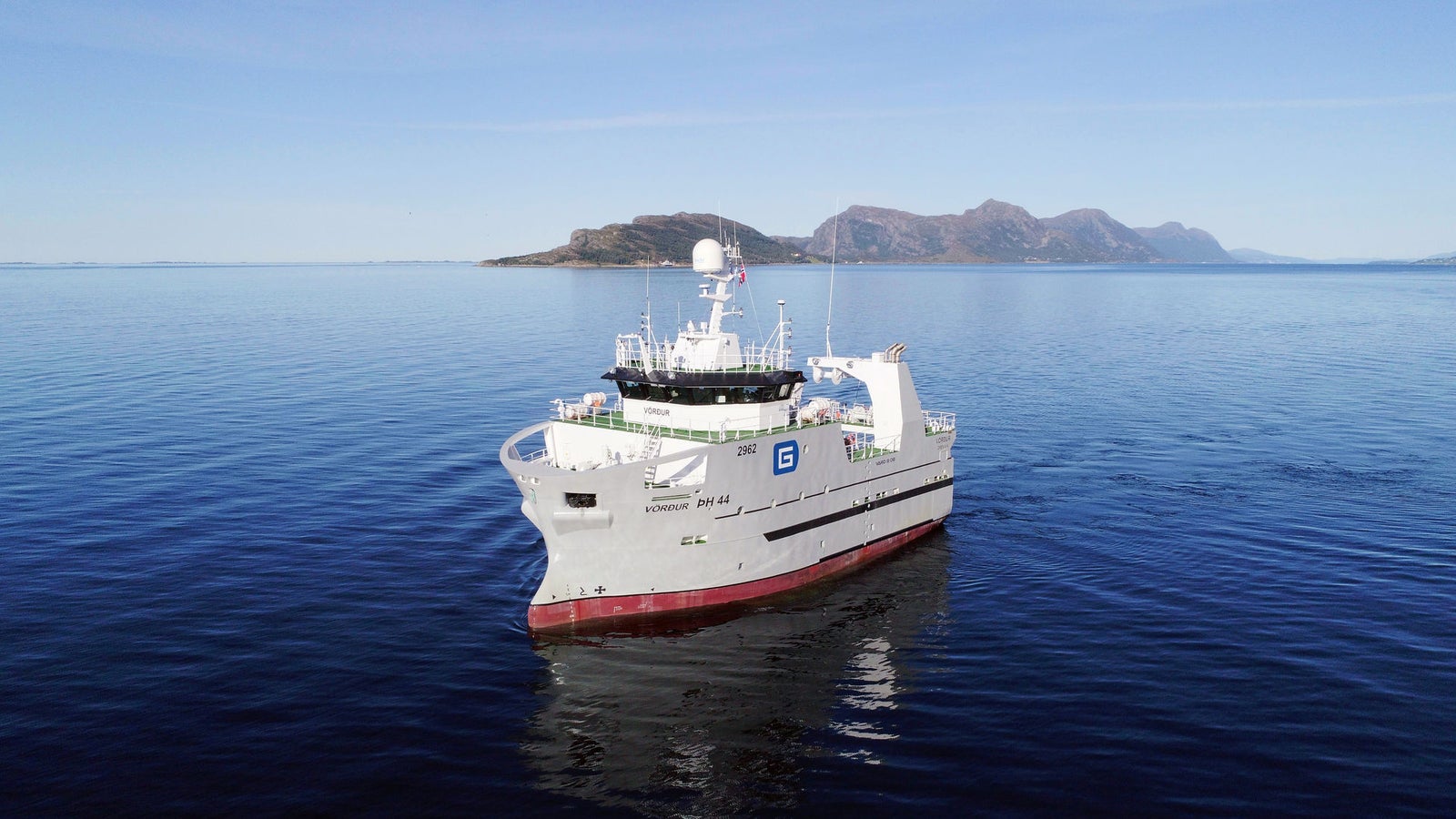 Prodotto - Nave da Ricerca Vordur: Vista laterale della nave operativa in navigazione su acque calme, scafo bianco con logo G visibile, attrezzature avanzate sul ponte, sfondo con isole e montagne distanti.