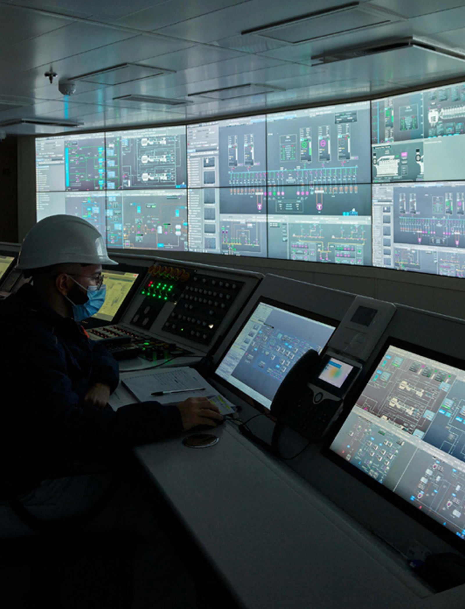  Fincantieri | Gruppo - Società - Controllate e collegate - HMSIT: Sala controllo moderna con persona con casco seduta alla postazione, molteplici schermi digitali mostrano interfacce di sistema e visualizzazioni dati complesse; ambiente high-tech per monitoraggio operativo o industriale, illuminazione soffusa che evidenzia la luminosità degli schermi.