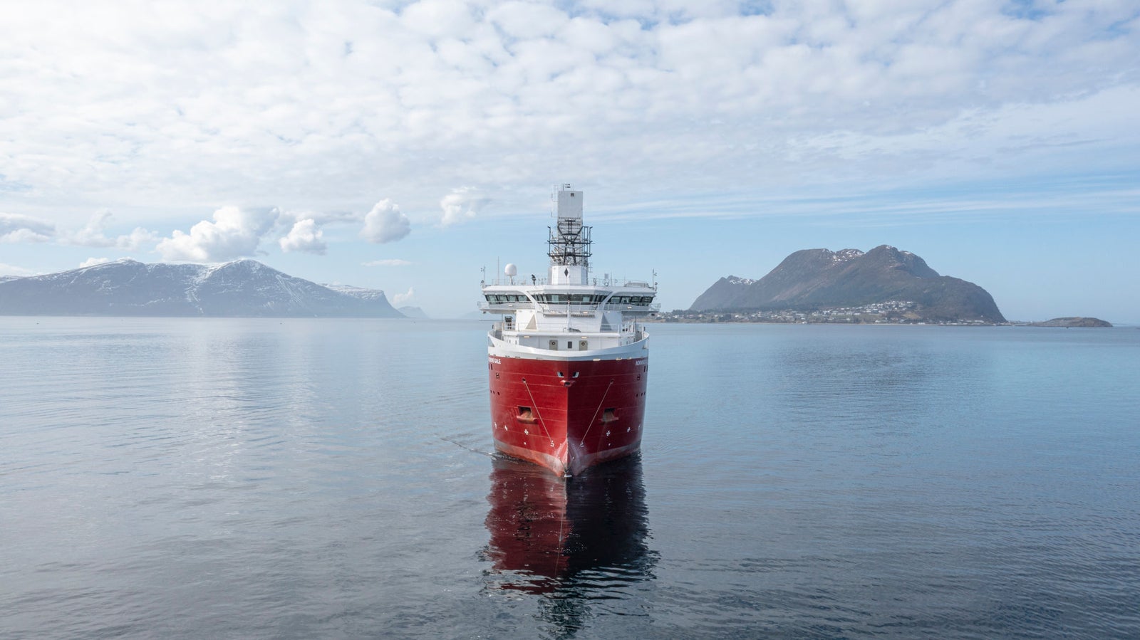 Fincantieri | Group - Subsidiaries and Affiliates - VARD: Striking red ship positioned centrally on serene waters, surrounded by Nordic landscape with mountains and cloudy skies; vessel features prominent tower structure and reflects on the still surface, scene evokes tranquility and showcases maritime engineering against natural backdrop.