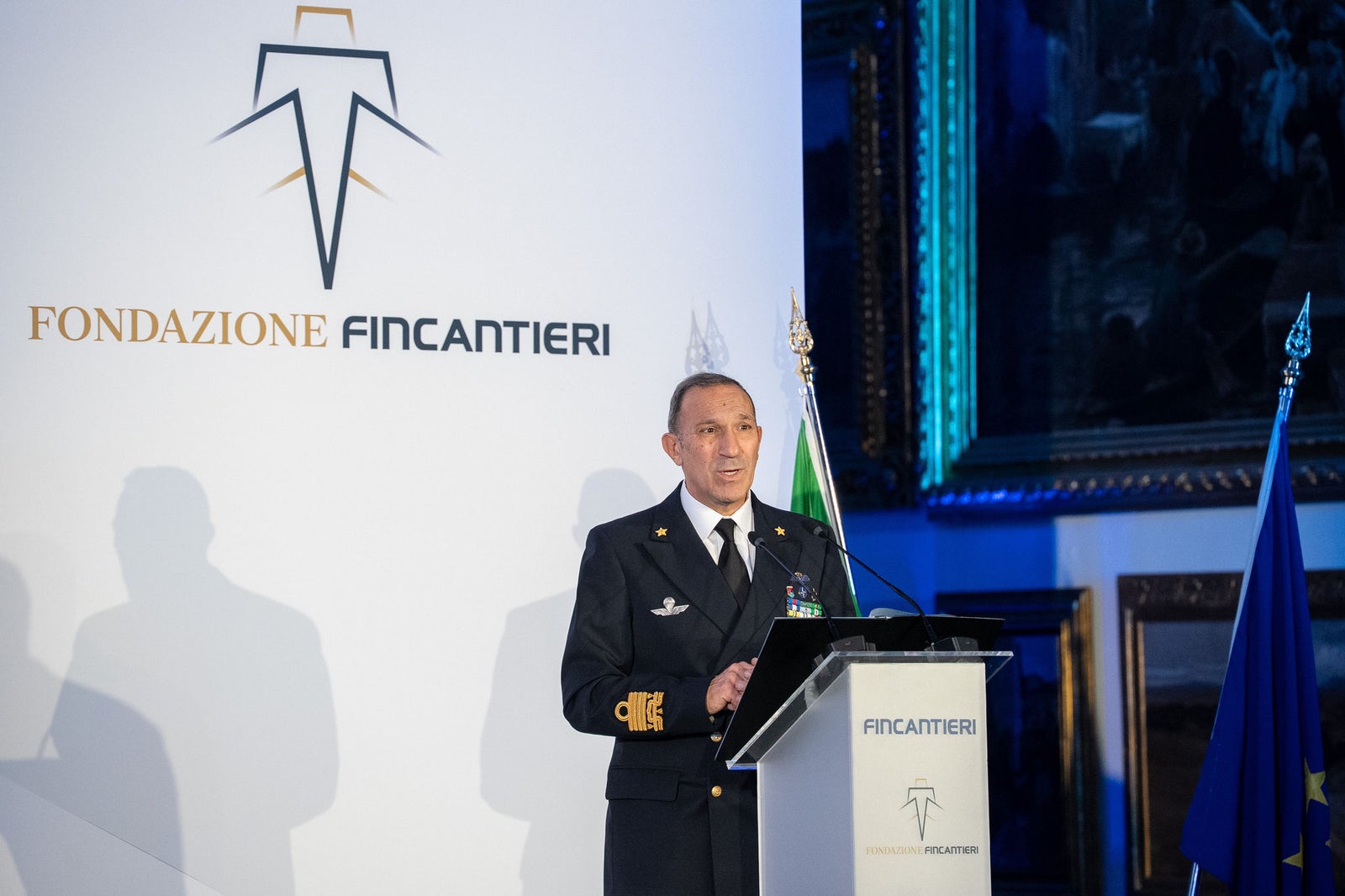Newsroom - Comunicati stampa - 2025 - Gruppo Fincantieri: Un anno di Fondazione Fincantieri: storia, innovazione e impatto sociale: Ufficiale alla Marina Militare interviene al podio durante l’evento della Fondazione Fincantieri, con logo istituzionale sullo sfondo e bandiere italiana ed europea a fianco.