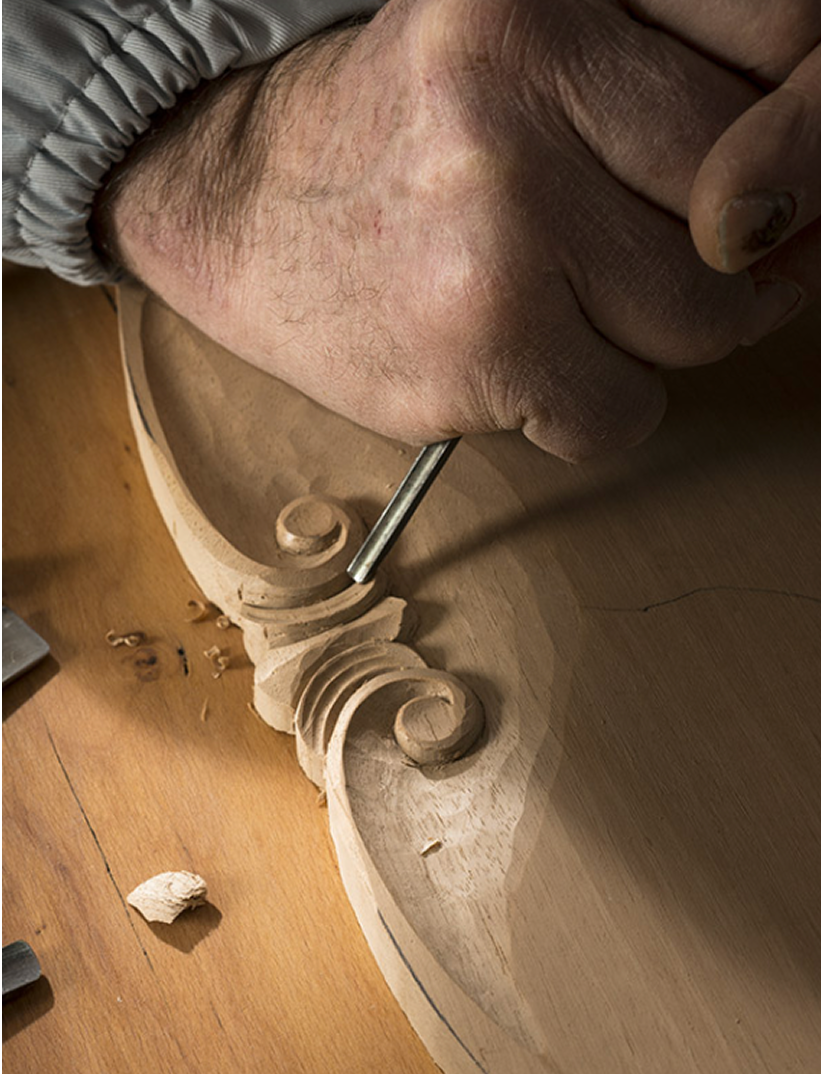 Fincantieri | Gruppo - Società - Controllate e collegate - Marine Interiors: Dettaglio di mano che intaglia motivi intricati su superficie in legno con scalpello, evidenza su maestria e precisione artigianale; trucioli visibili e texture del legno, focus su tecniche di lavorazione tradizionale.